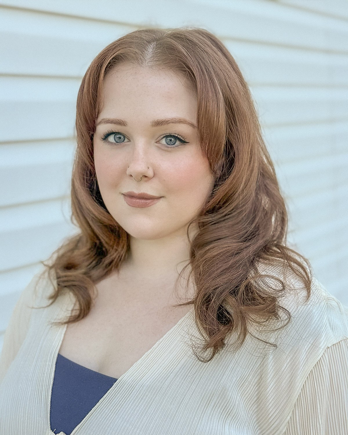Young woman with wavy brown hair and blue eyes, wearing a light-colored top, standing outdoors near a white wall.