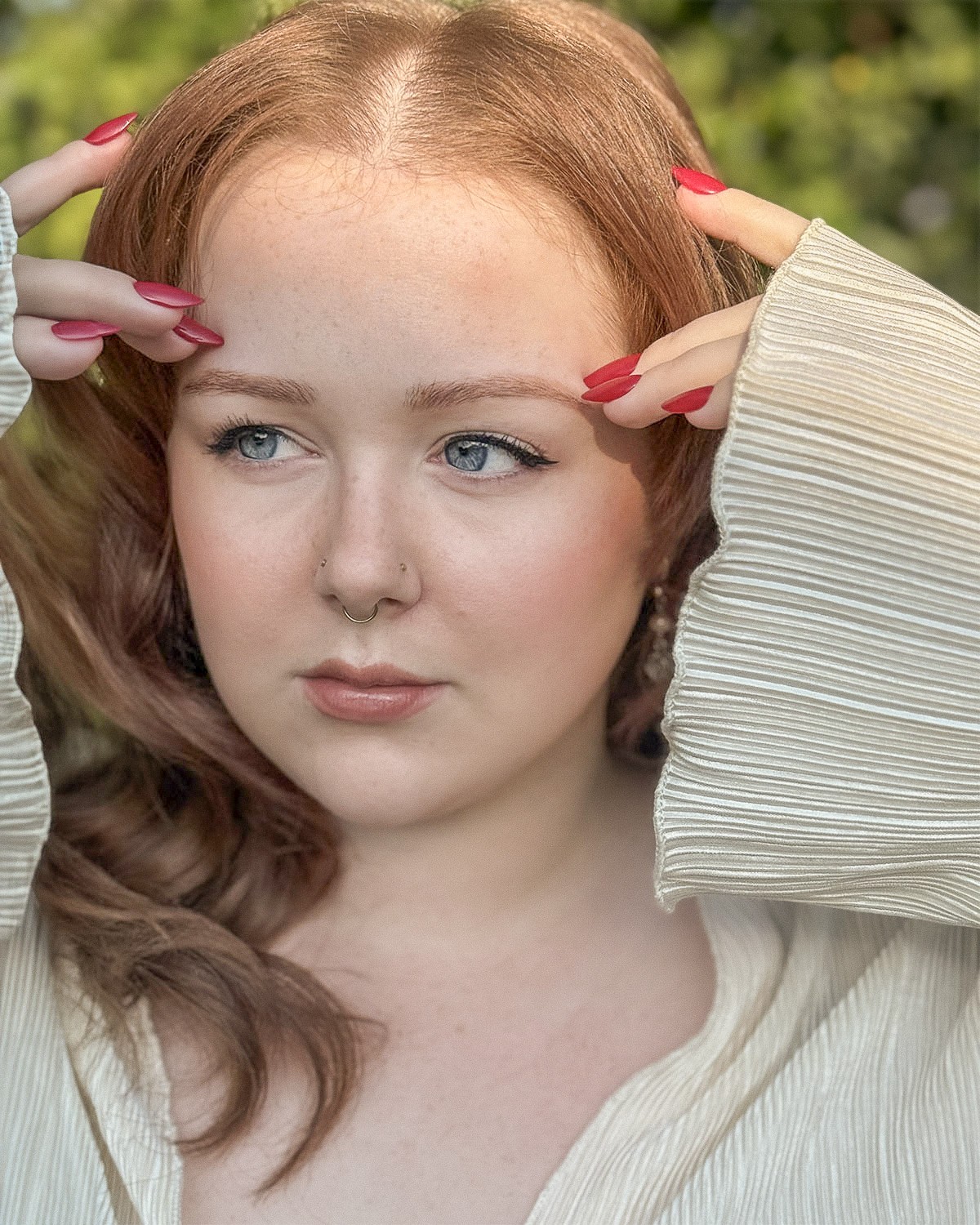 Young woman with red hair and blue eyes, touching her forehead with both hands, outdoors with greenery background.
