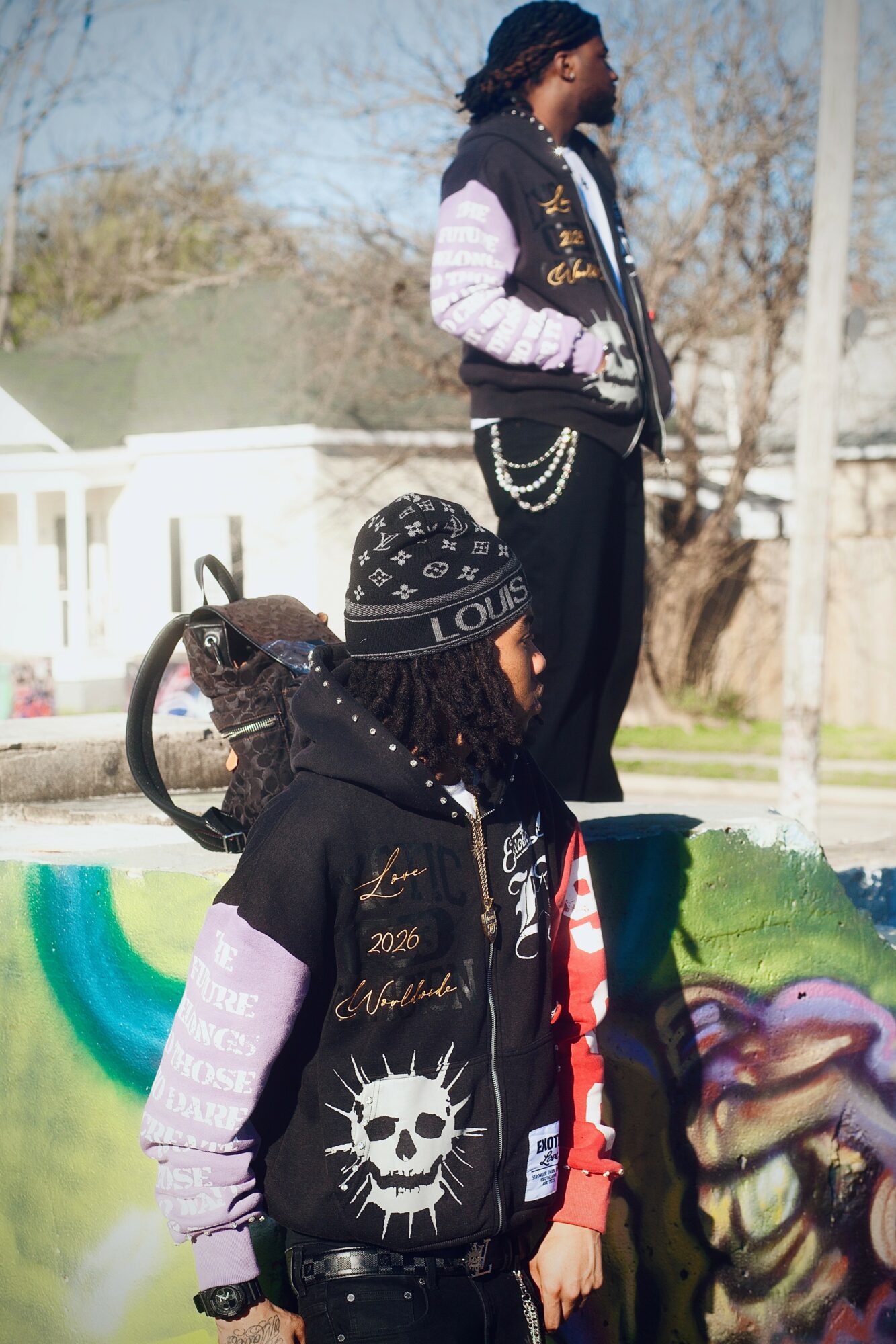 Two young people outdoors, one standing and one sitting, with colorful graffiti and trees in the background.