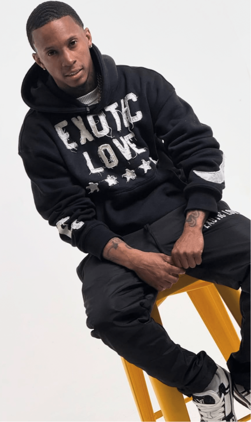 Young man sitting on a yellow stool, wearing a black hoodie with white text, black pants, and sneakers, against a white background.