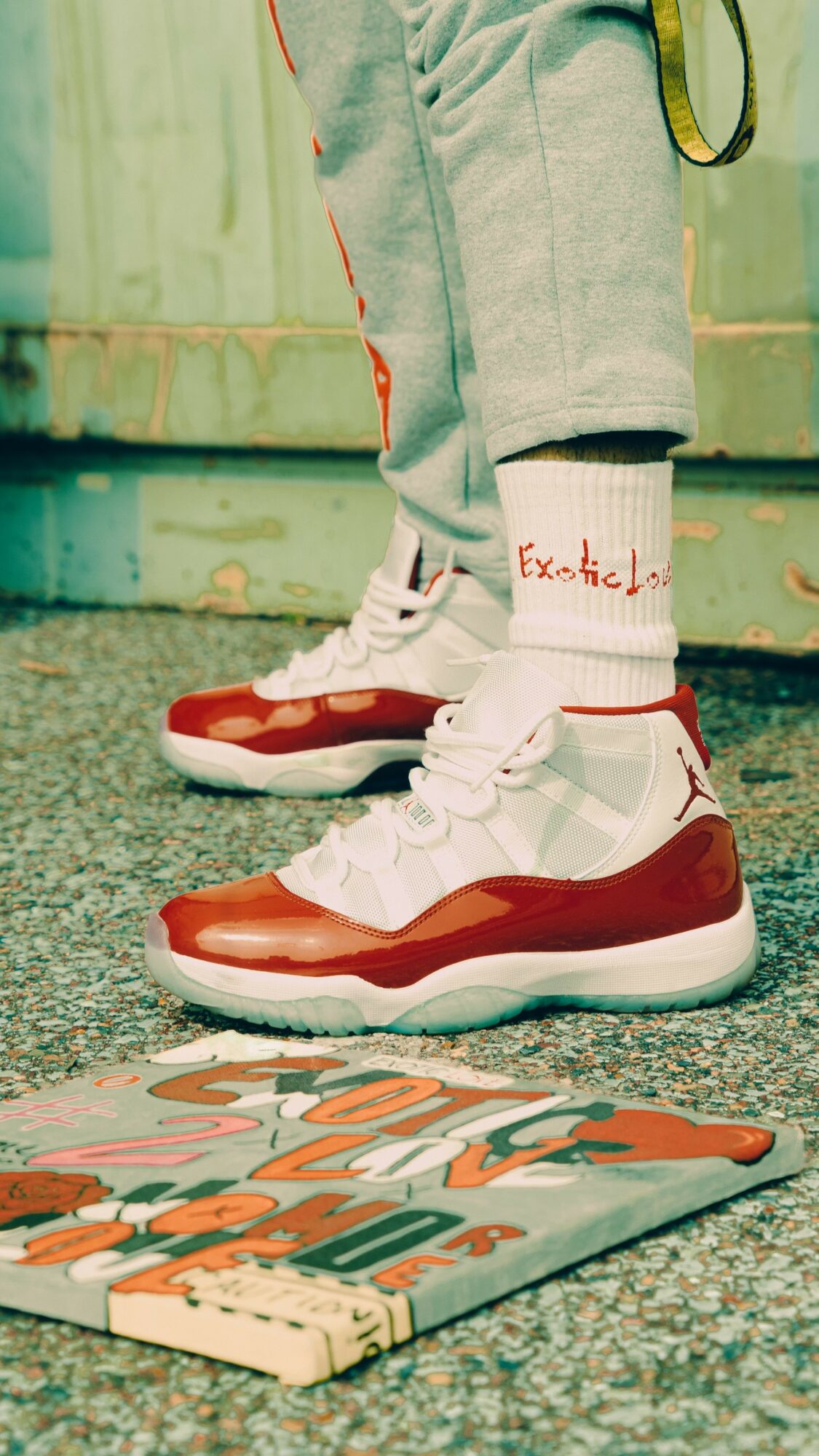 Person wearing white and red sneakers, gray sweatpants, and white socks with red text, standing on a textured ground near a colorful skateboard.