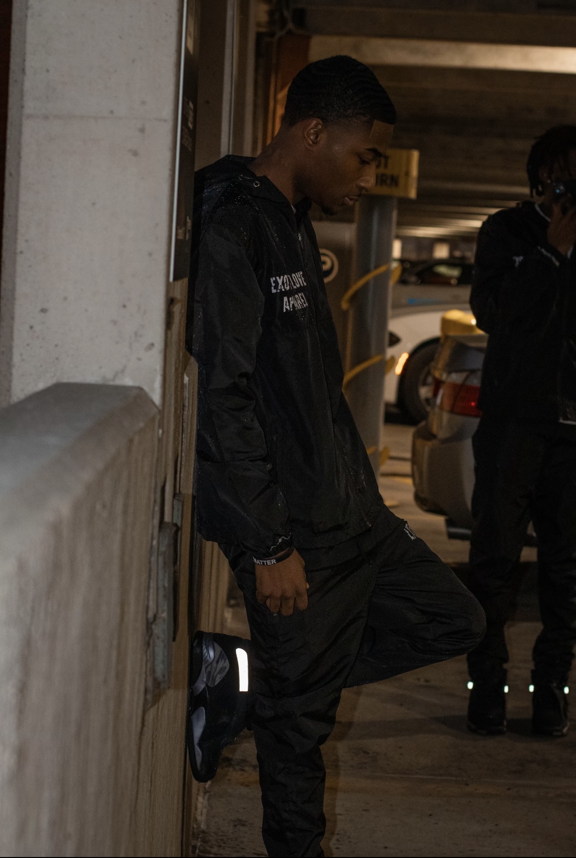 Young man leaning against a wall in an indoor parking area, wearing a black jacket and pants, with cars in the background.