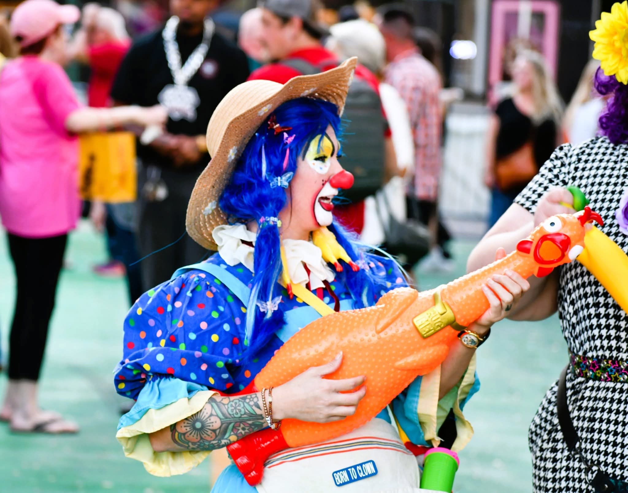 Clown with blue hair, colorful costume, holding a large orange toy, in a crowded outdoor setting.