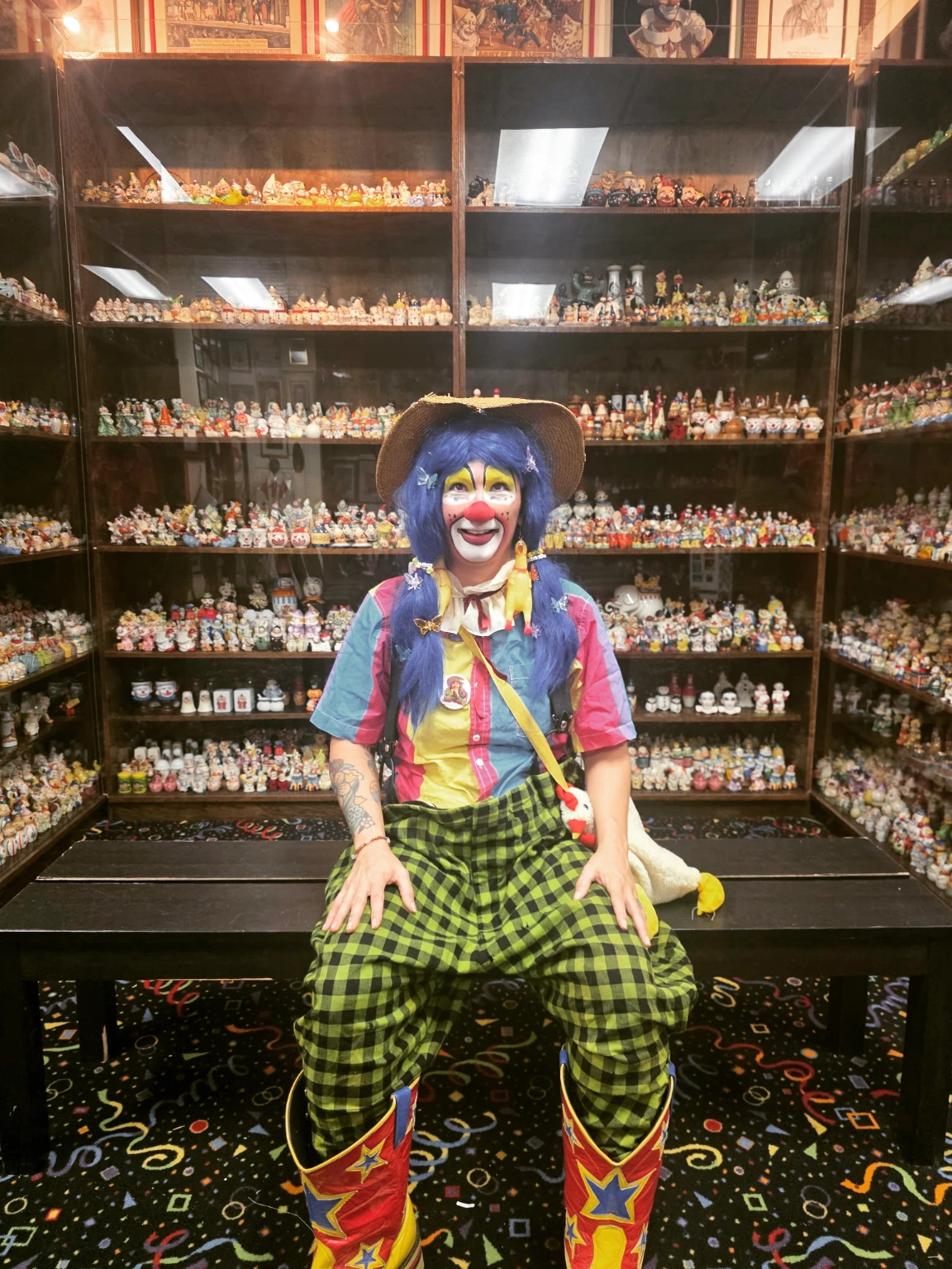 Clown figure with colorful costume, hat, and makeup, sitting in front of shelves filled with miniature figurines.
