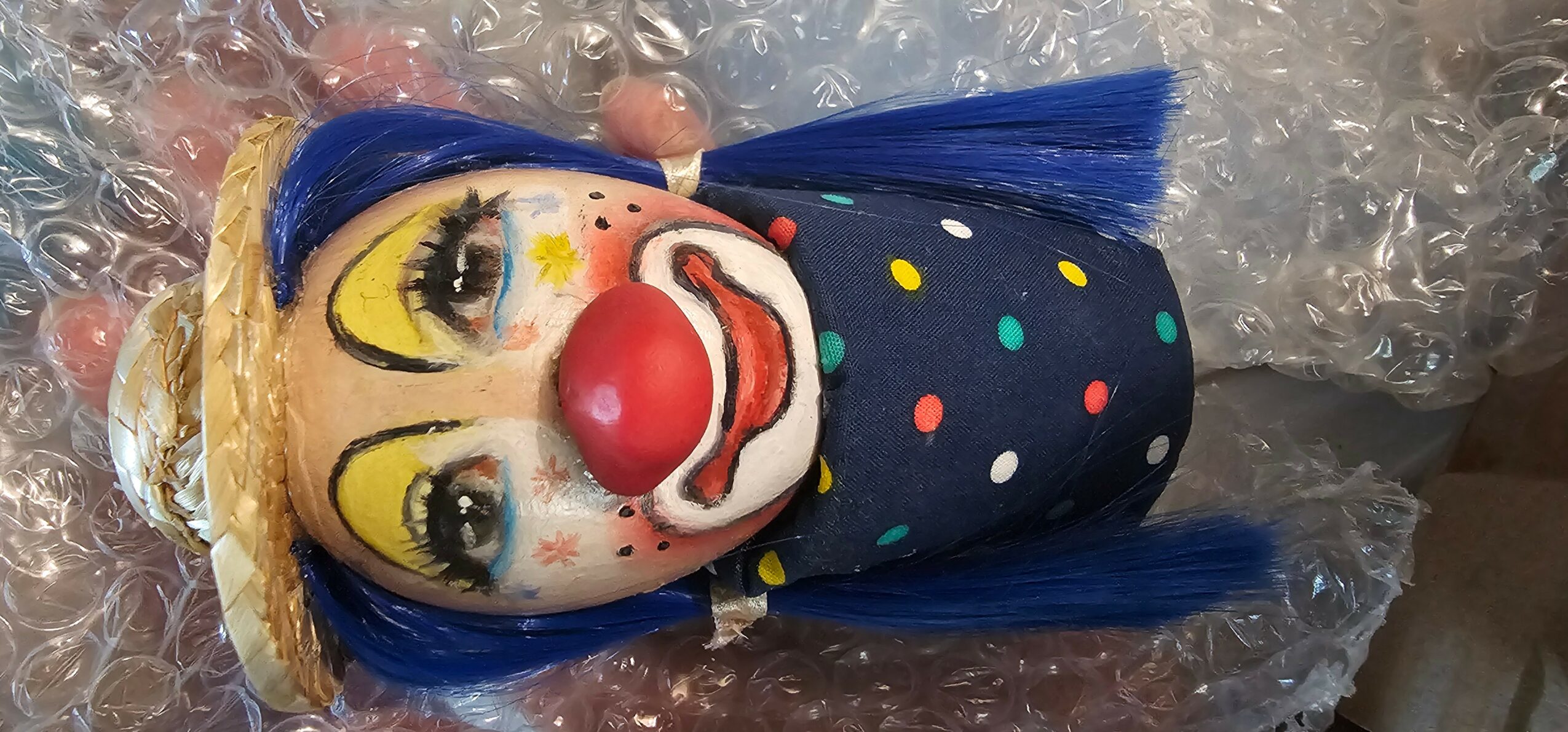 Clown doll with blue hair, yellow eyes, red nose, and polka-dot outfit, lying on bubble wrap.