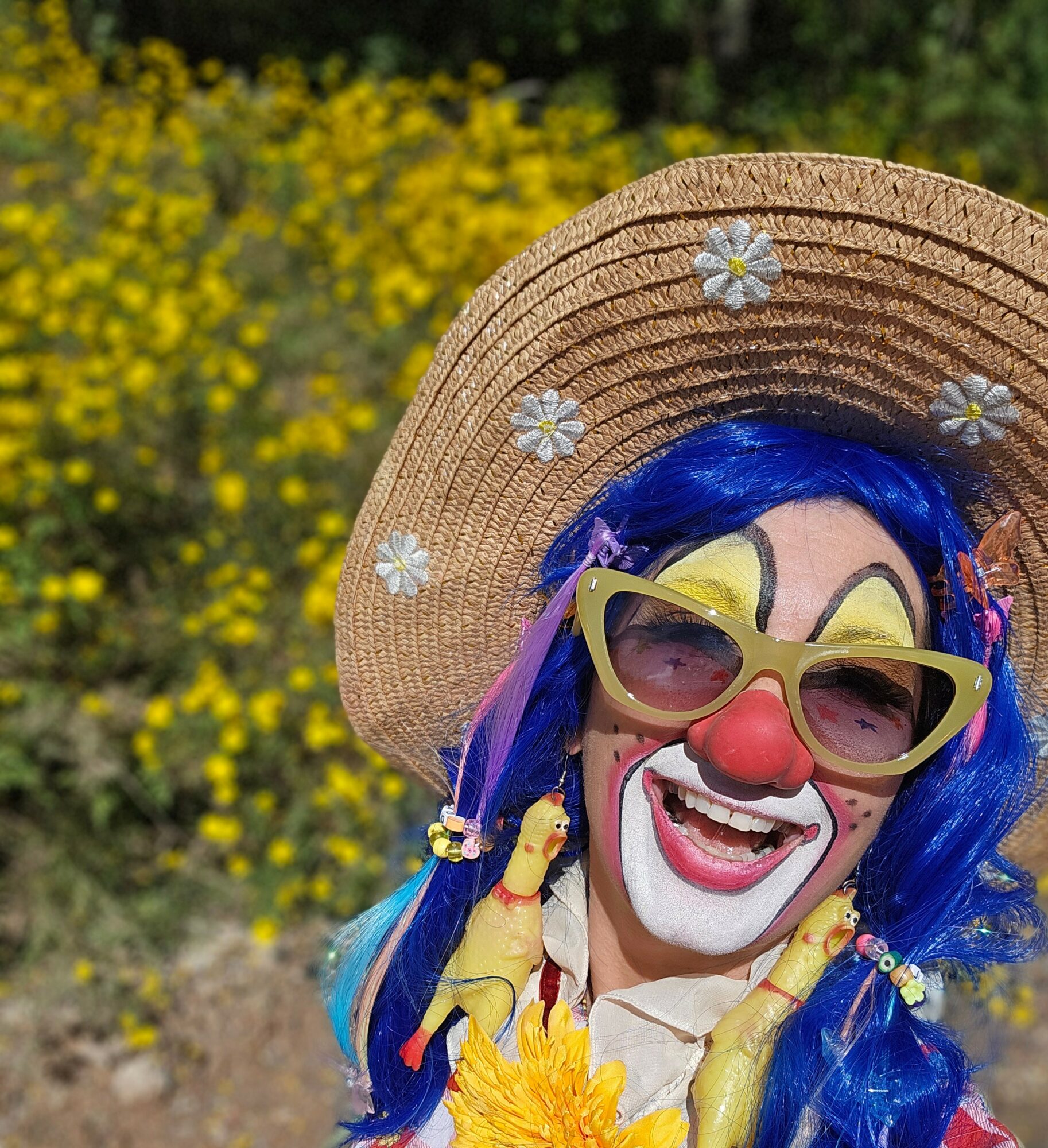 Person dressed as a clown with blue hair, yellow glasses, and a straw hat, smiling outdoors with yellow flowers in background.