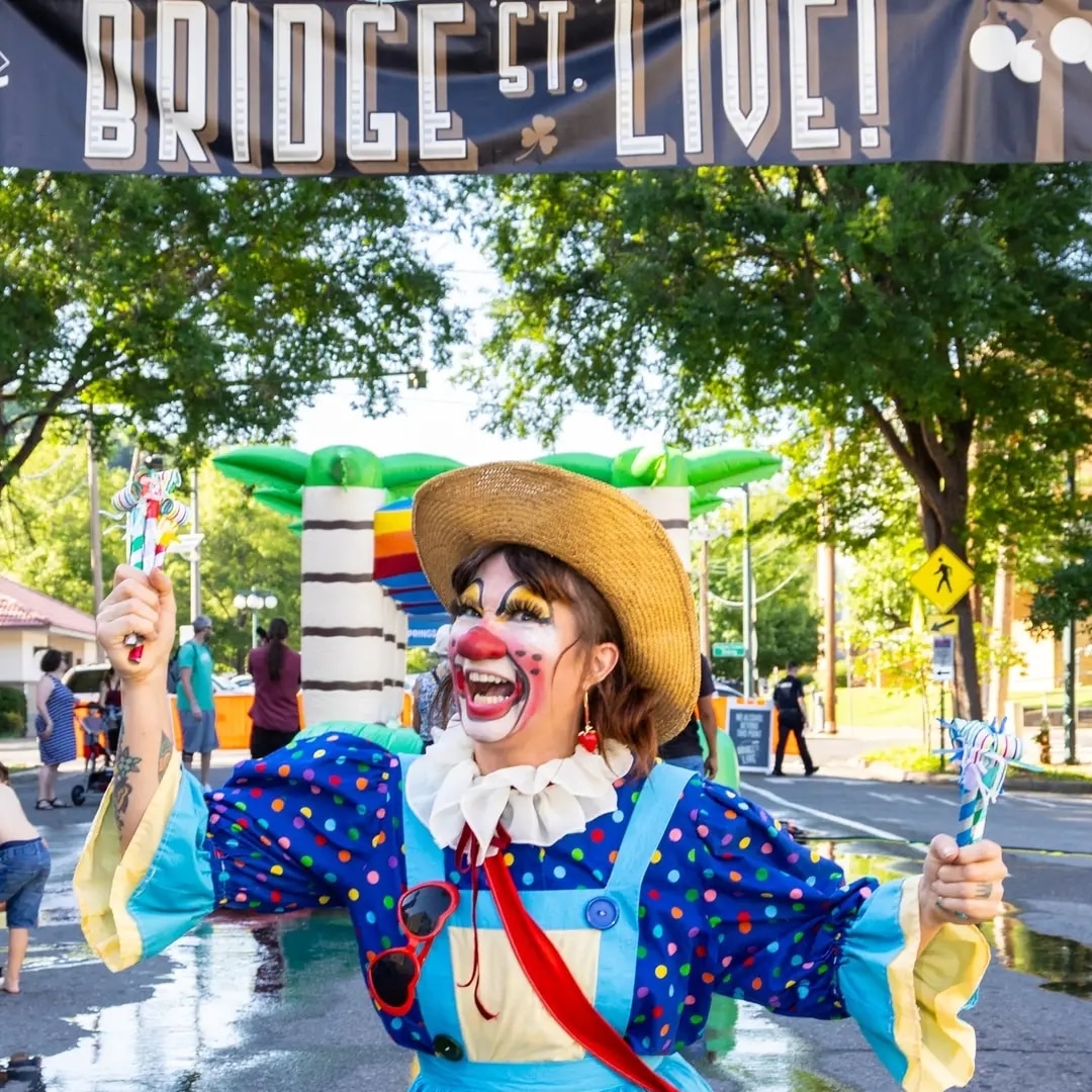 Clown in colorful costume and hat smiling, holding party poppers outdoors with trees and people in background.