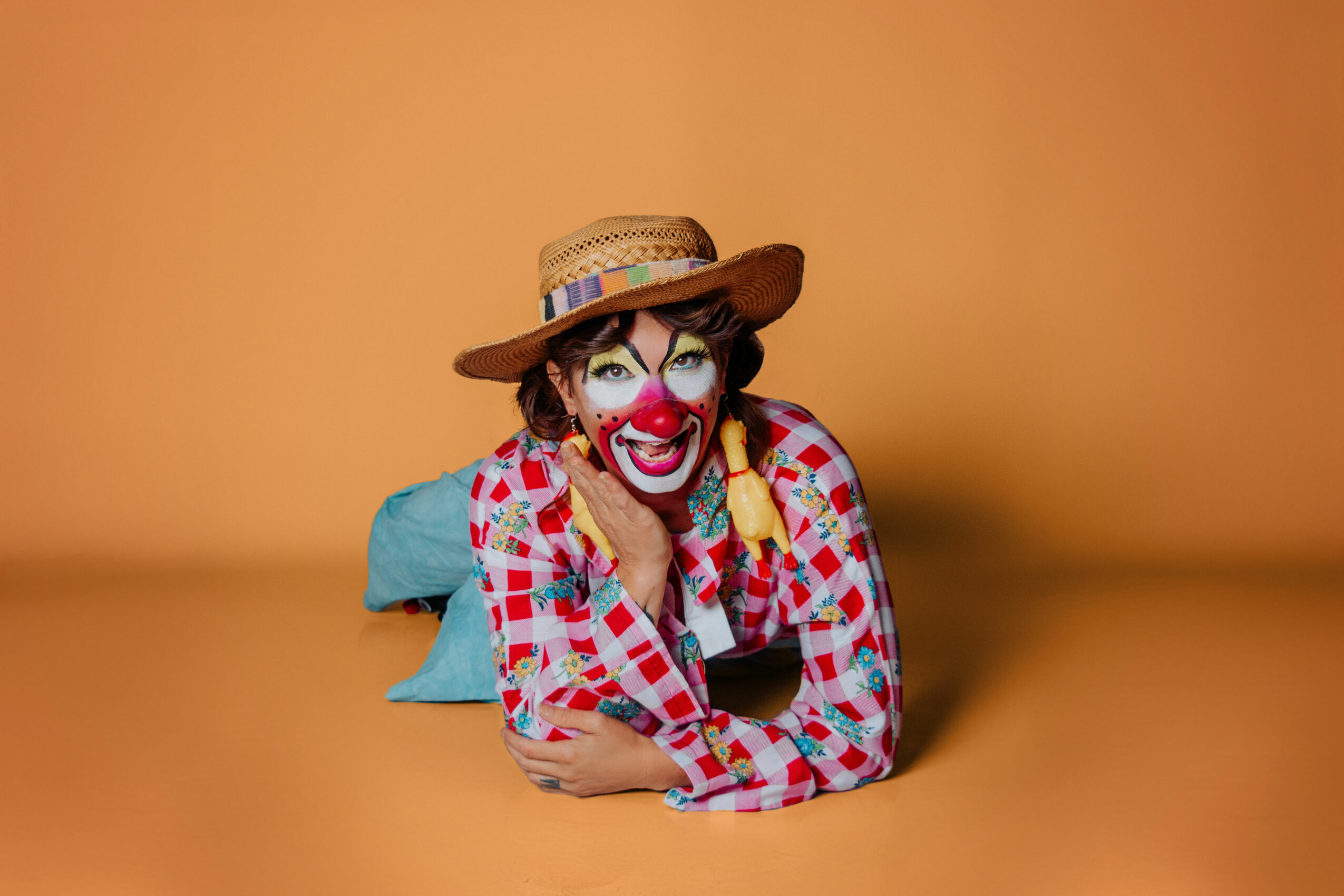 Person with clown makeup, wearing a straw hat and checkered shirt, lying on a blue pillow against an orange background.