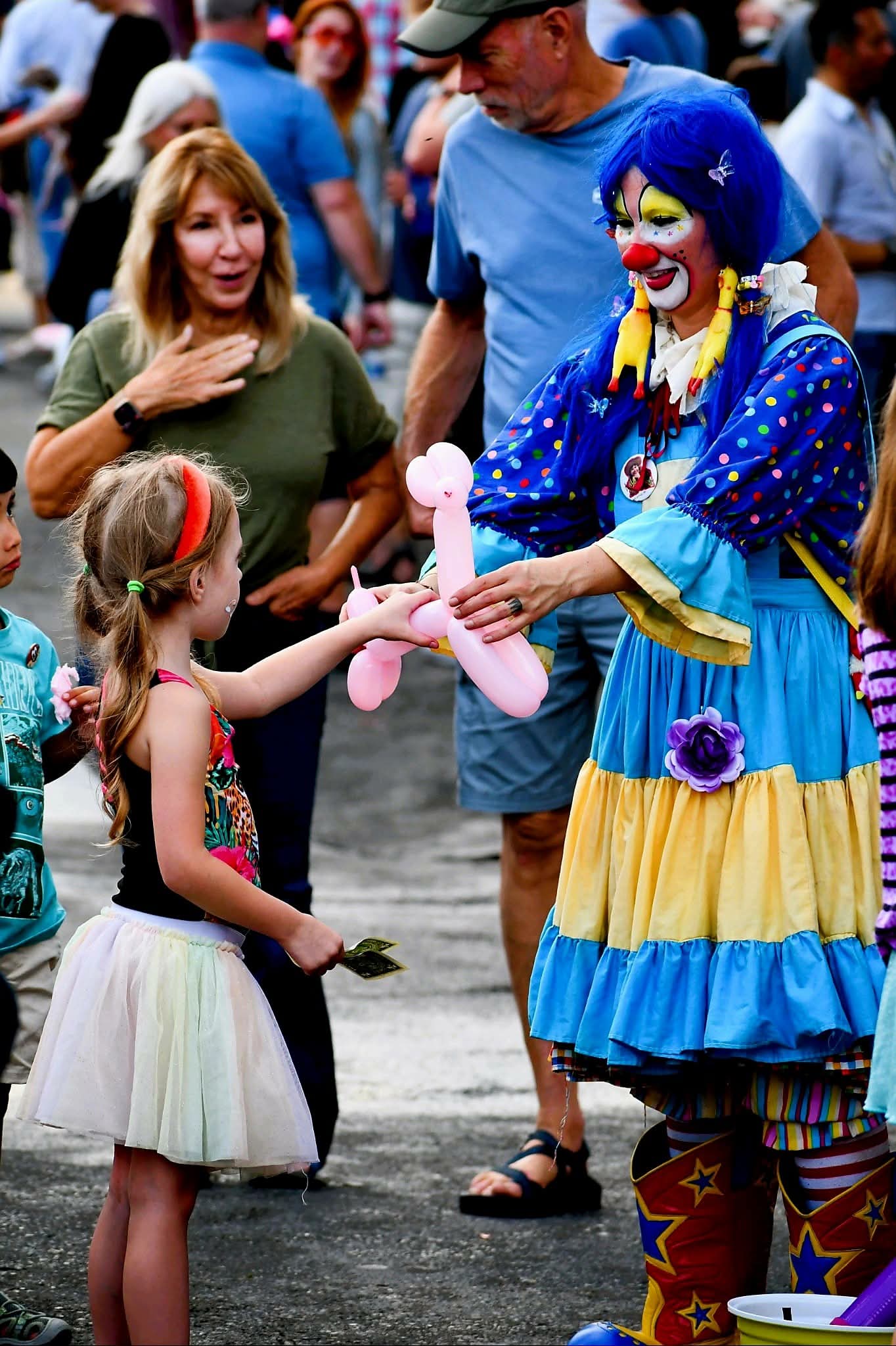 Clown in colorful costume hands a balloon animal to a young girl at a crowded outdoor event.