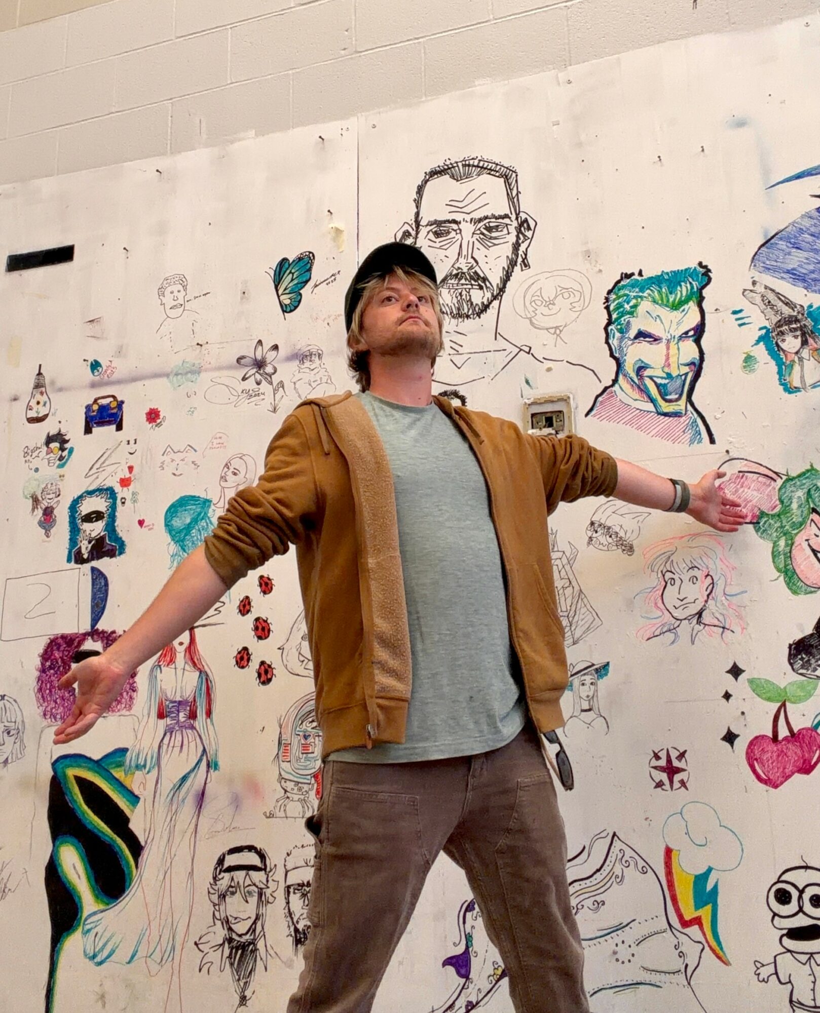 Person with arms outstretched standing in front of a wall with colorful drawings and sketches, including faces and abstract designs.
