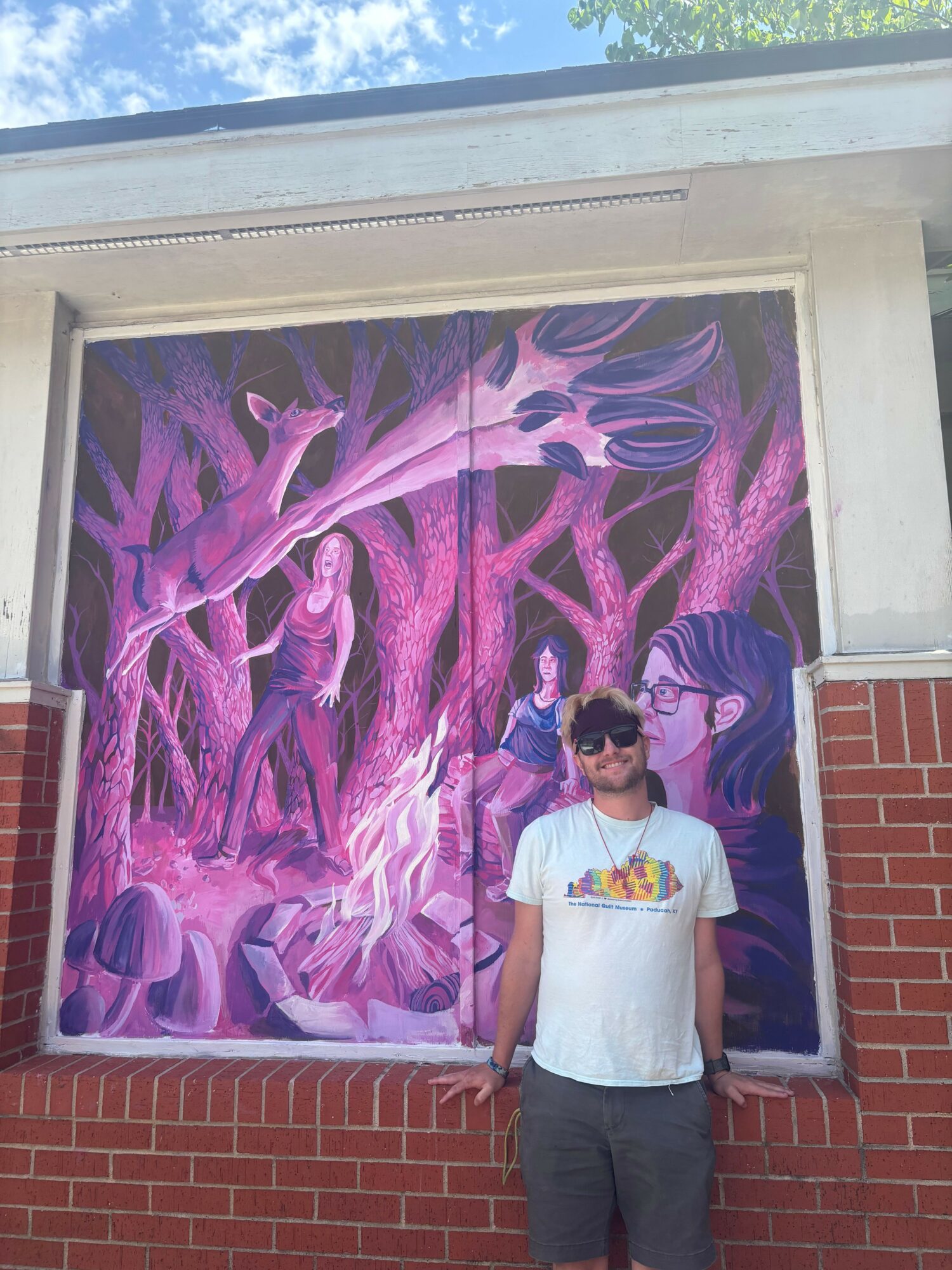 Person standing next to a colorful, abstract mural with purple and pink hues, on a brick wall background.