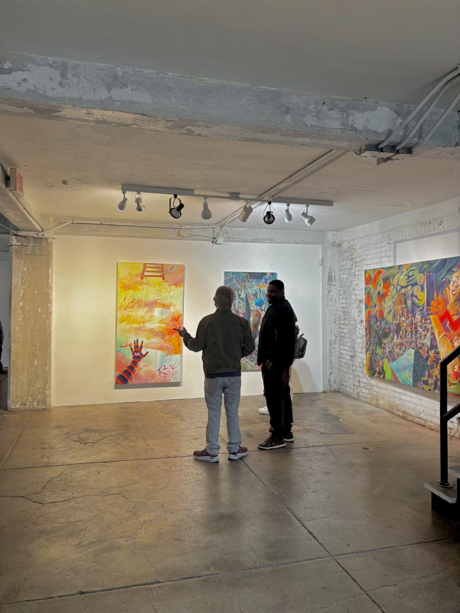 Two people stand in an art gallery, viewing colorful abstract paintings on the wall.