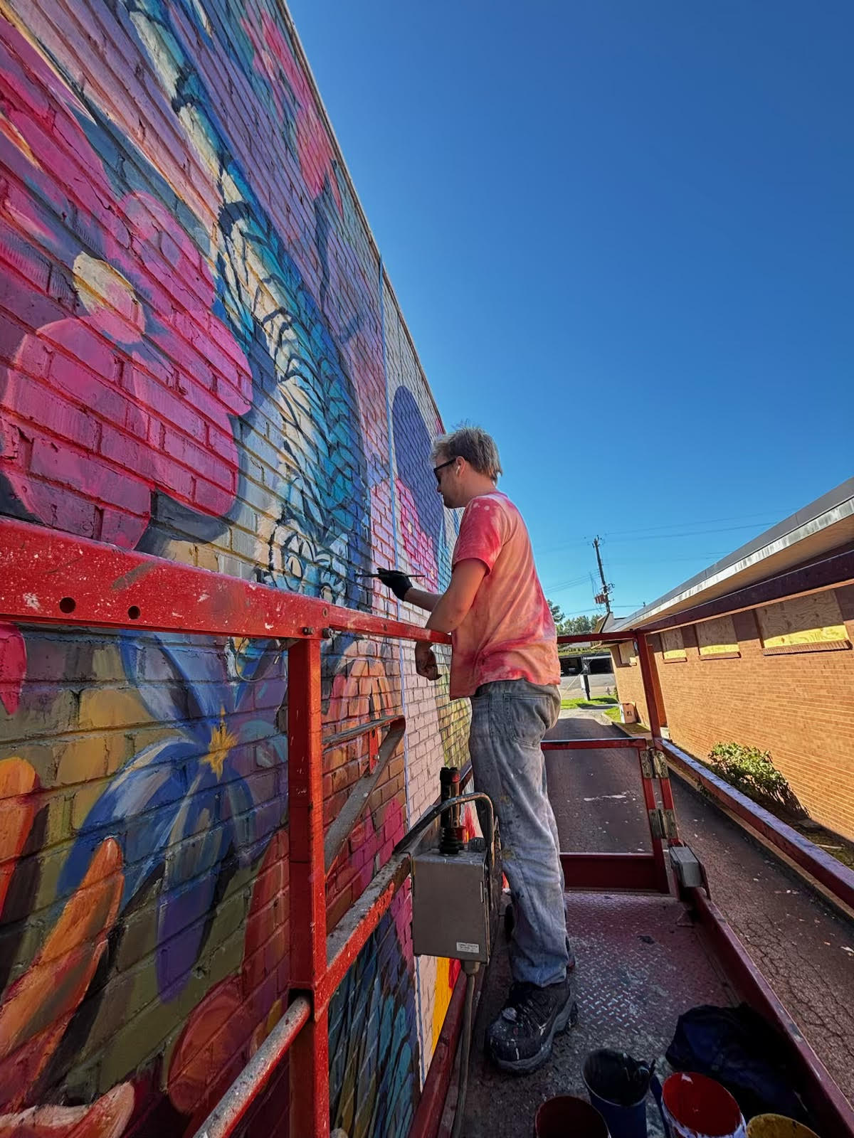 Person painting colorful graffiti on brick wall, standing on scaffolding, with blue sky background.