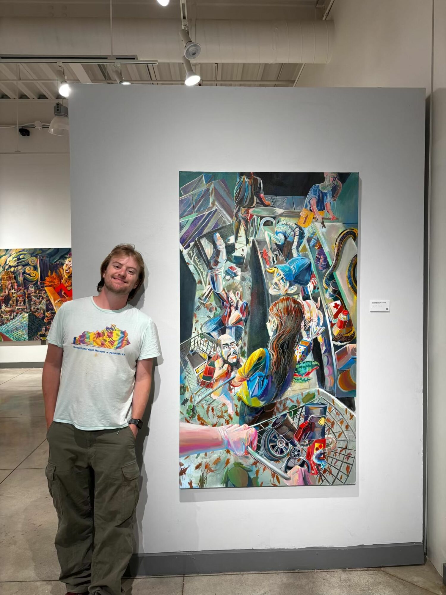 Person standing in front of colorful abstract artwork on gallery wall, smiling, wearing a white T-shirt and dark pants.
