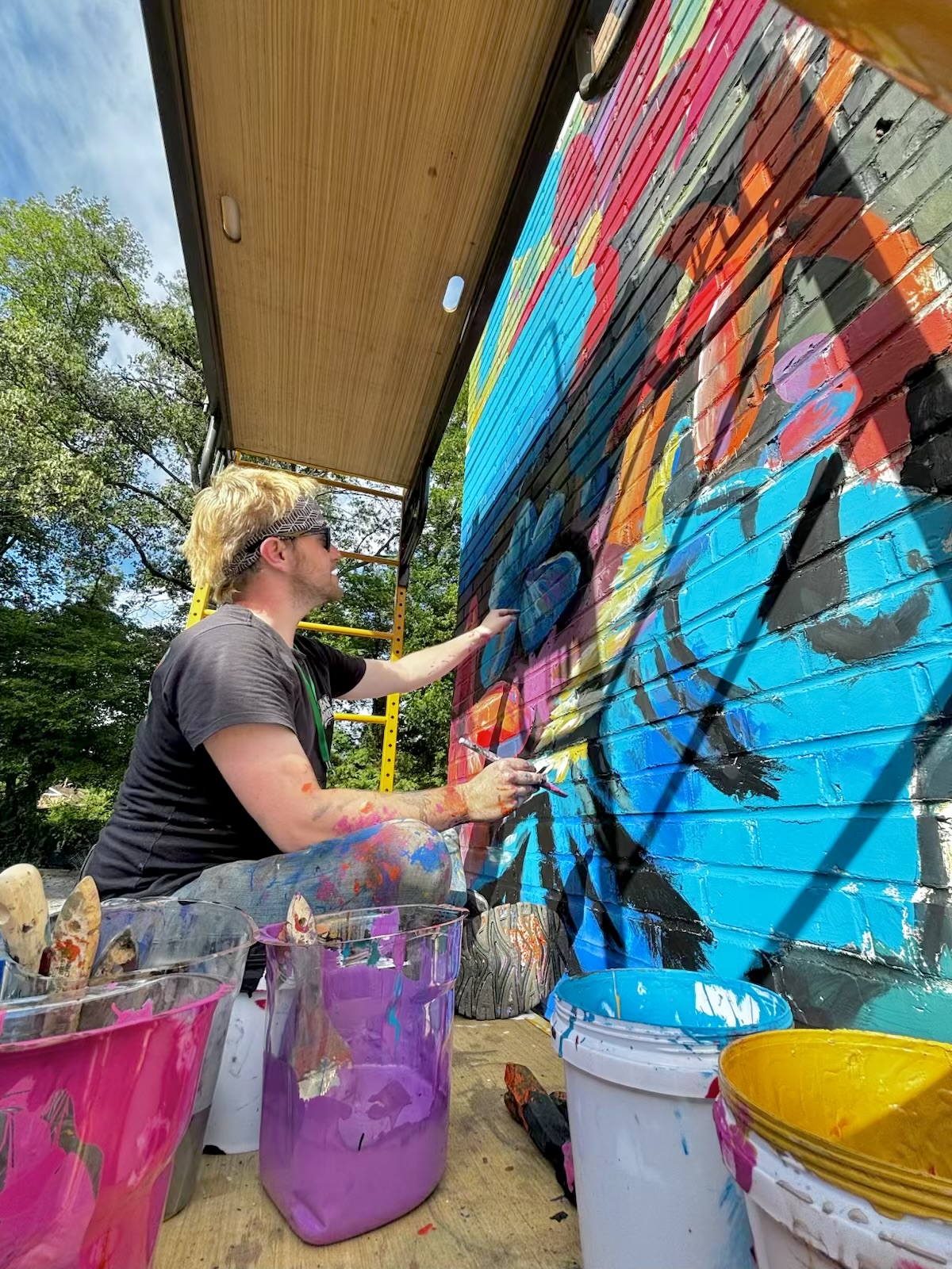Person painting a colorful mural on a wall outdoors, with paint buckets nearby, trees in the background.