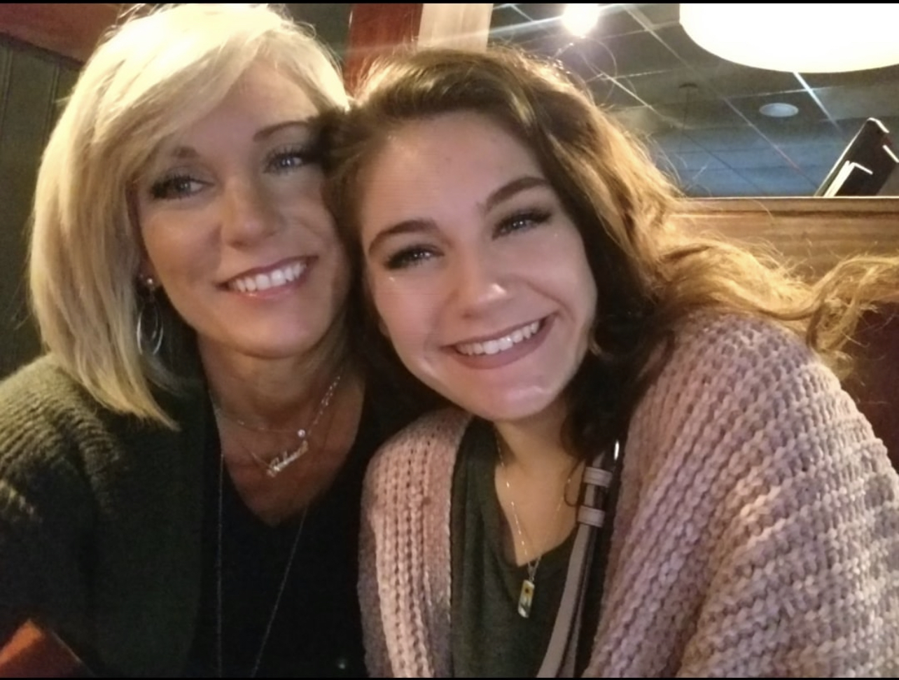 Two women smiling closely, one with blonde hair and the other with brown hair, in a cozy indoor setting.