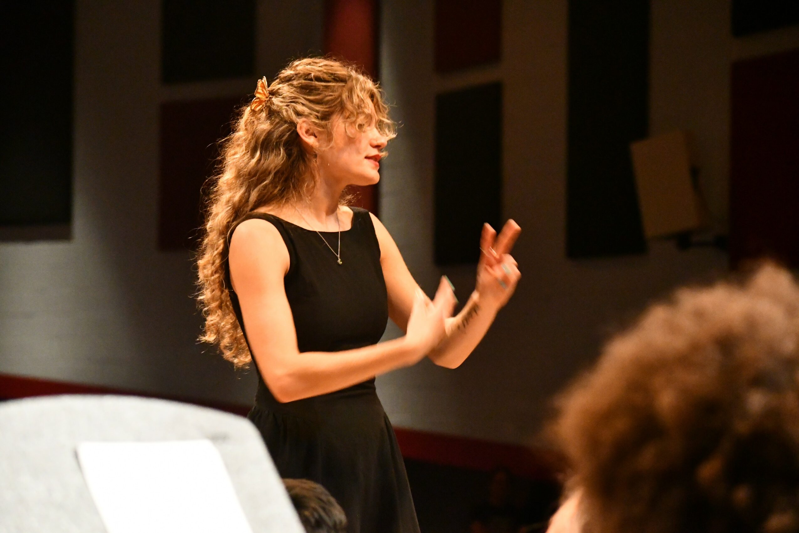 Woman with curly hair in a black dress gesturing with hands during a performance or presentation.