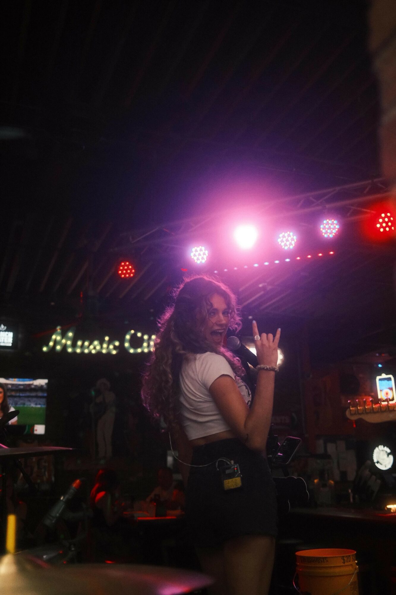 Person with curly hair in a white top and dark shorts standing in a dimly lit venue with colorful stage lights, music equipment, and a sign reading 'Music'.