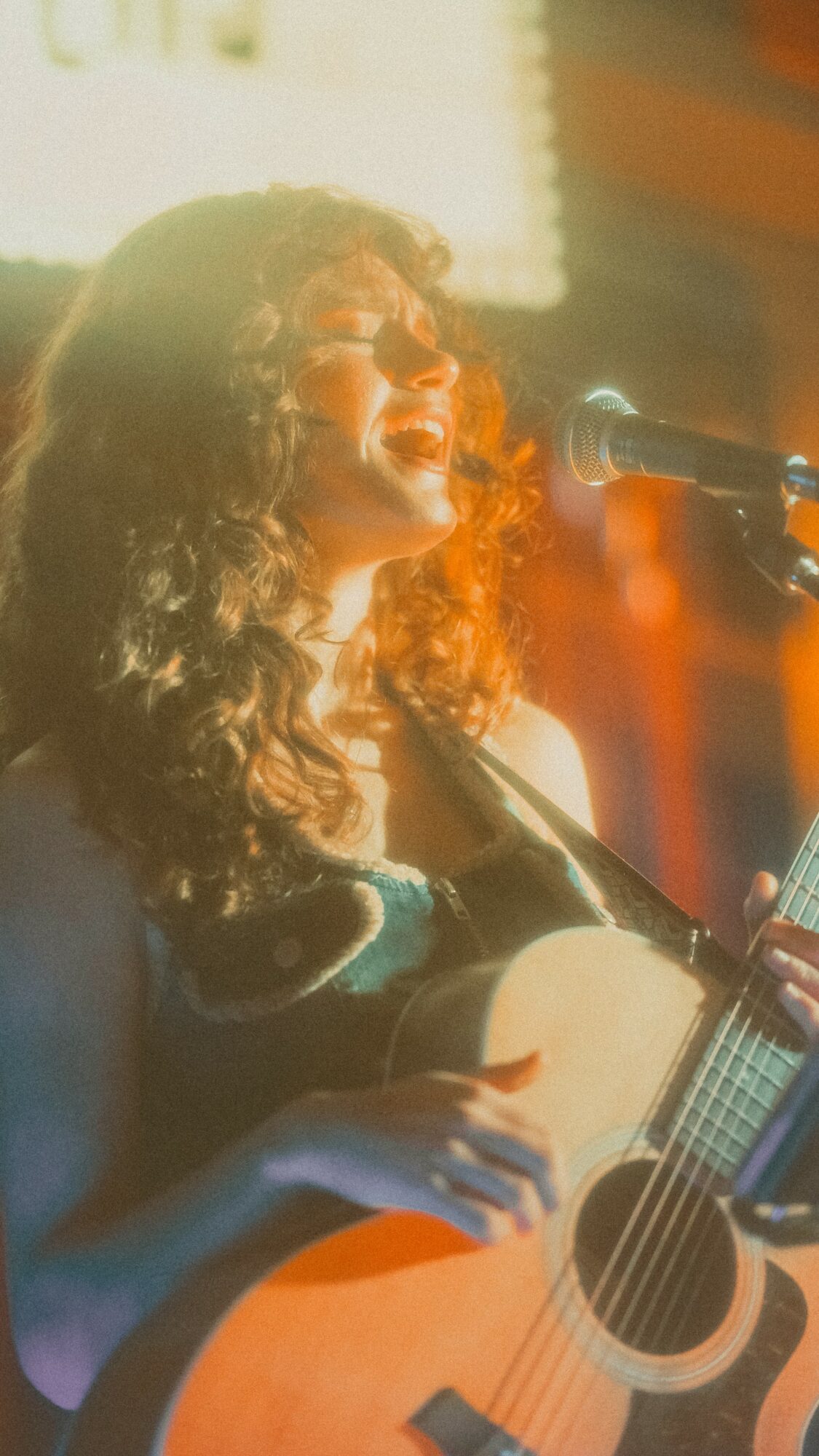 Person with curly hair singing into a microphone while playing an acoustic guitar, stage lighting in background.