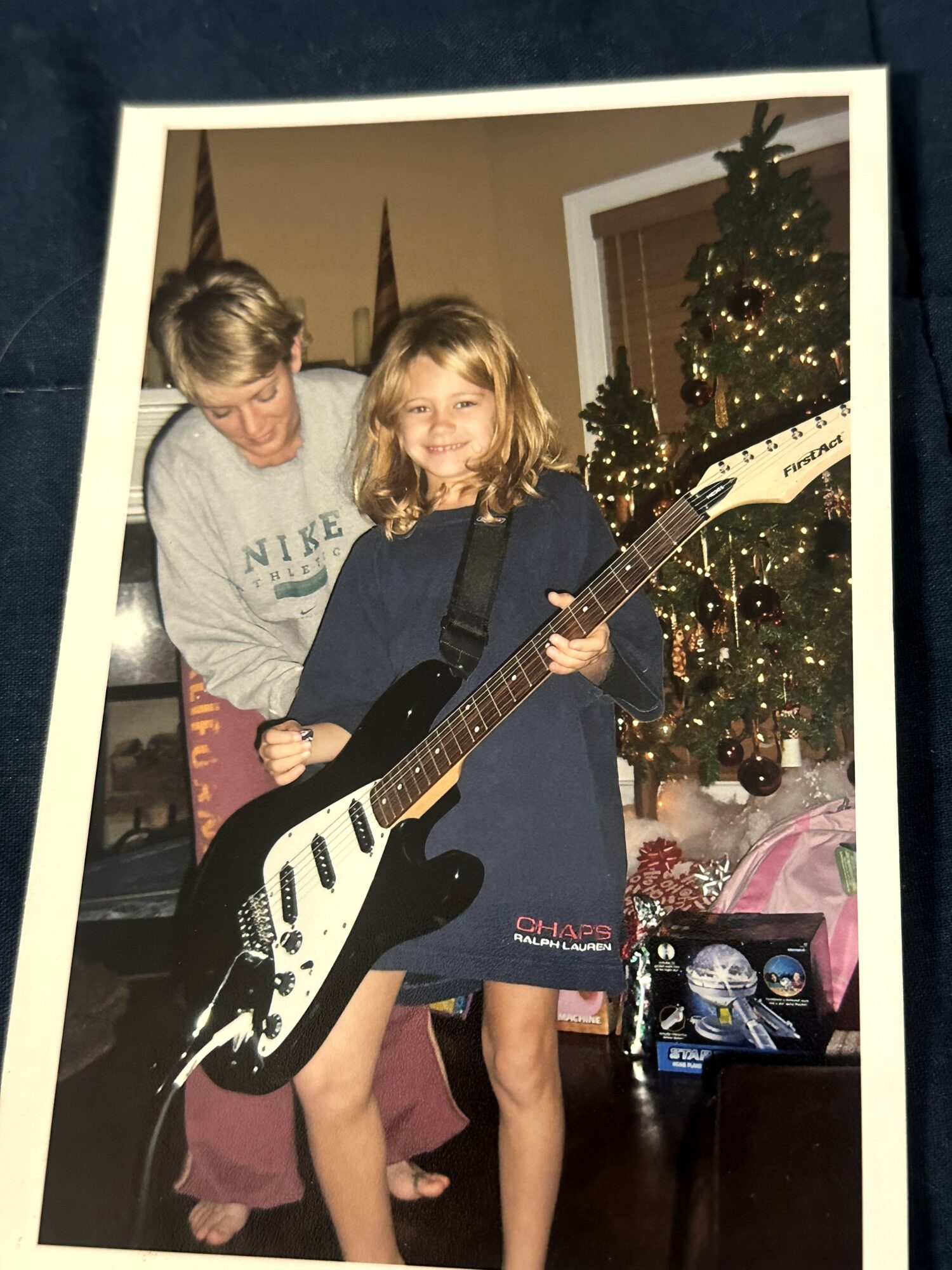 A girl holding a guitar with an adult behind her, Christmas tree in the background, in a decorated room.