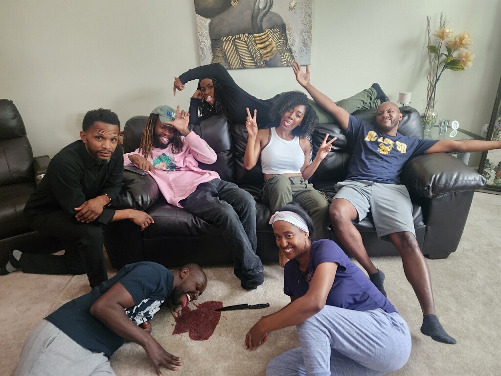 Six people sitting and lying on a black sofa in a living room, with a painting and a plant in the background.