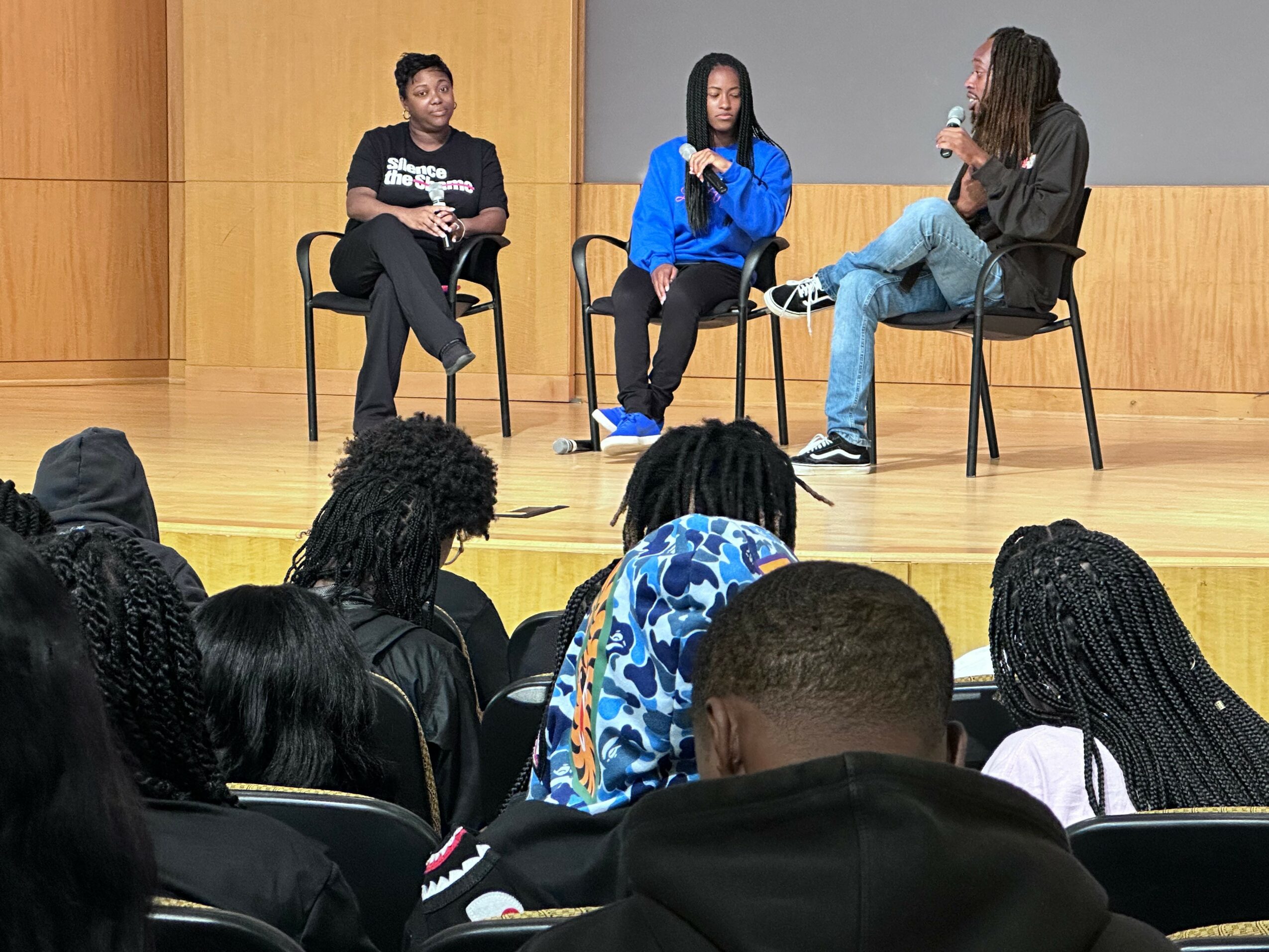 Three people sit on stage in chairs, engaging in a discussion, with an audience watching attentively.