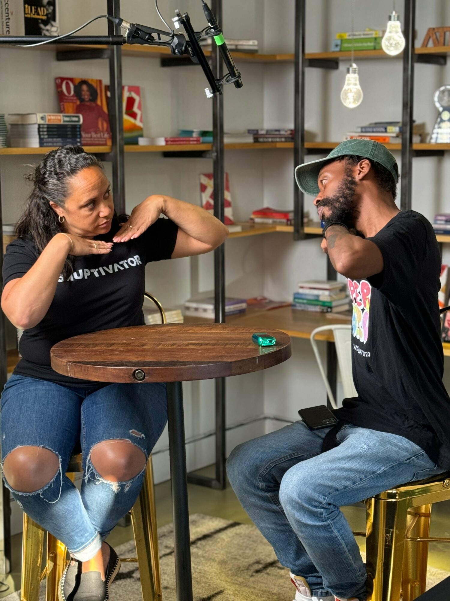Two people sit at a round table in a room with shelves and hanging light bulbs, engaging in conversation.