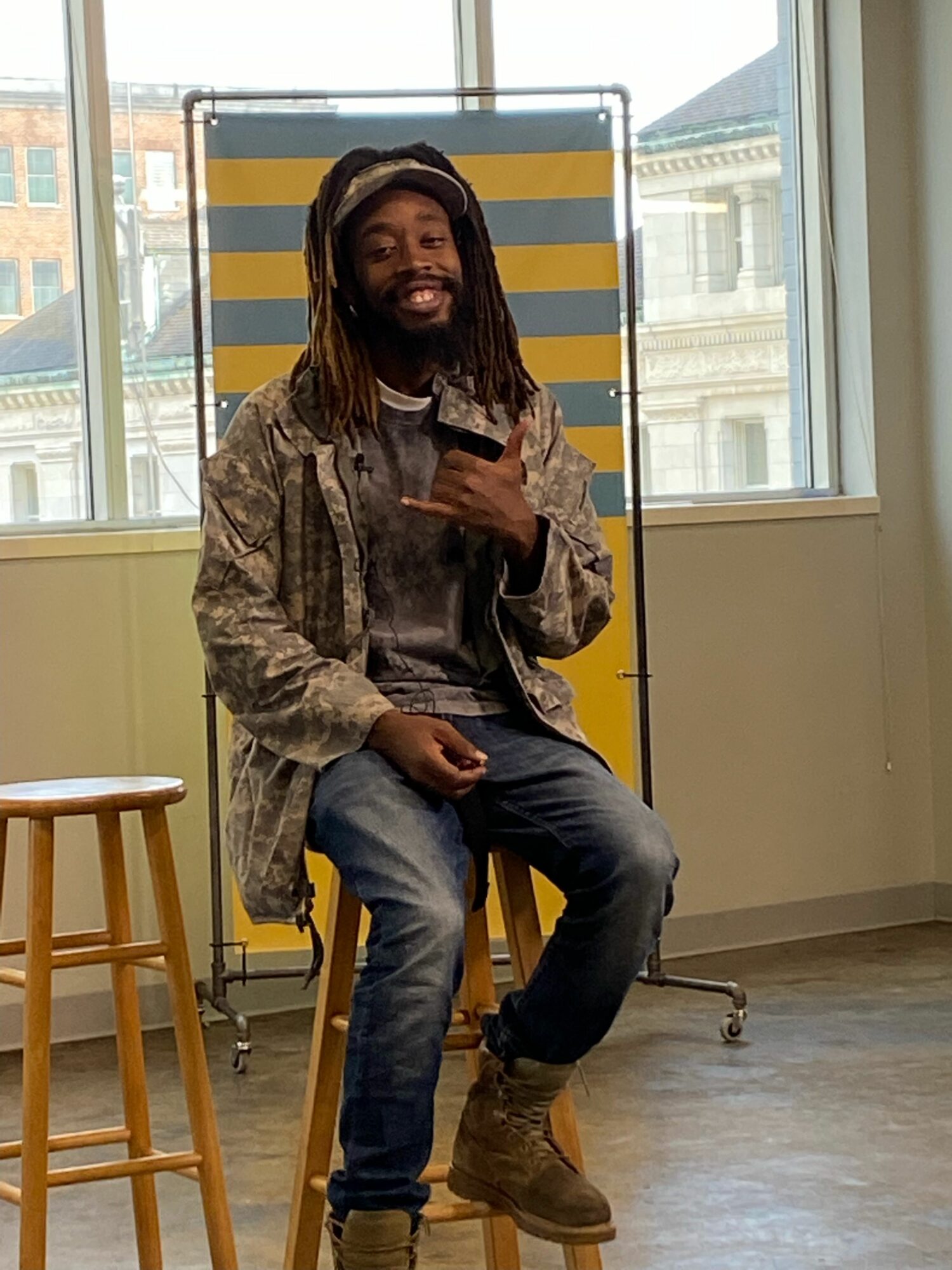 Person standing indoors near large window, holding a phone, with a yellow and gray striped backdrop behind them.