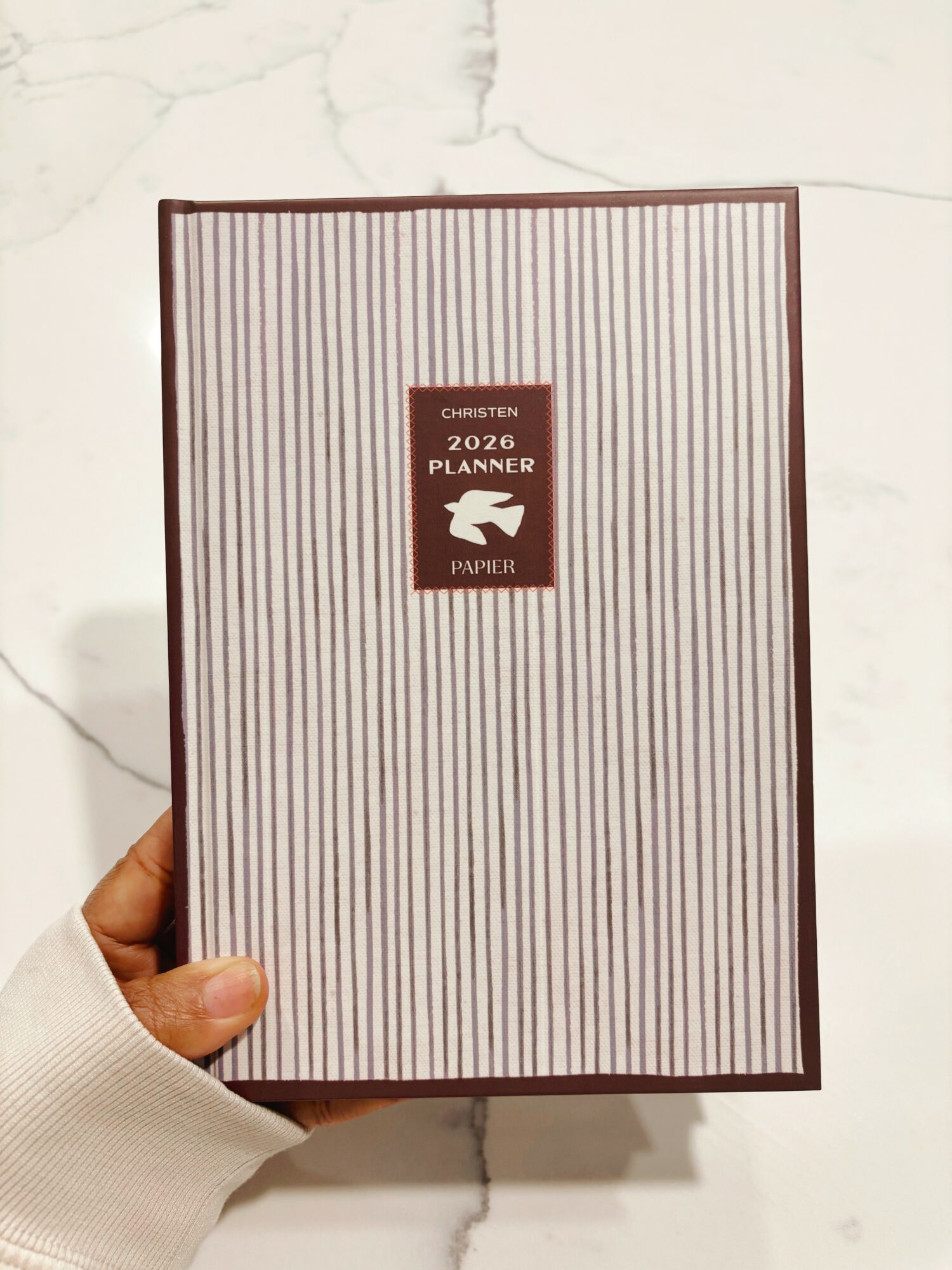 Person holding a 2020 planner with vertical striped cover, label in the center, against a white marble background.
