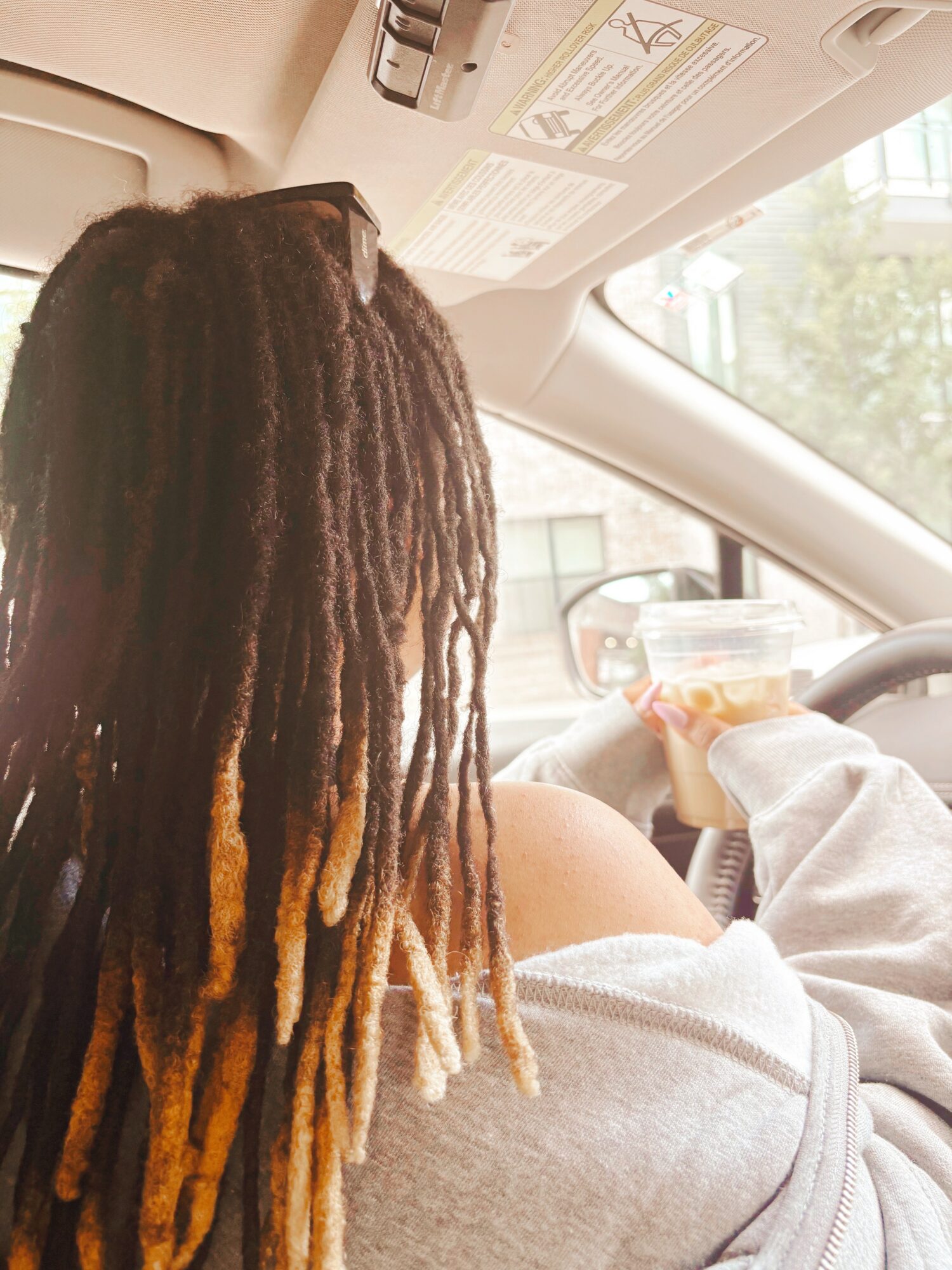 Person with dreadlocks sitting in car, holding a drink, wearing a light-colored hoodie, view from behind.