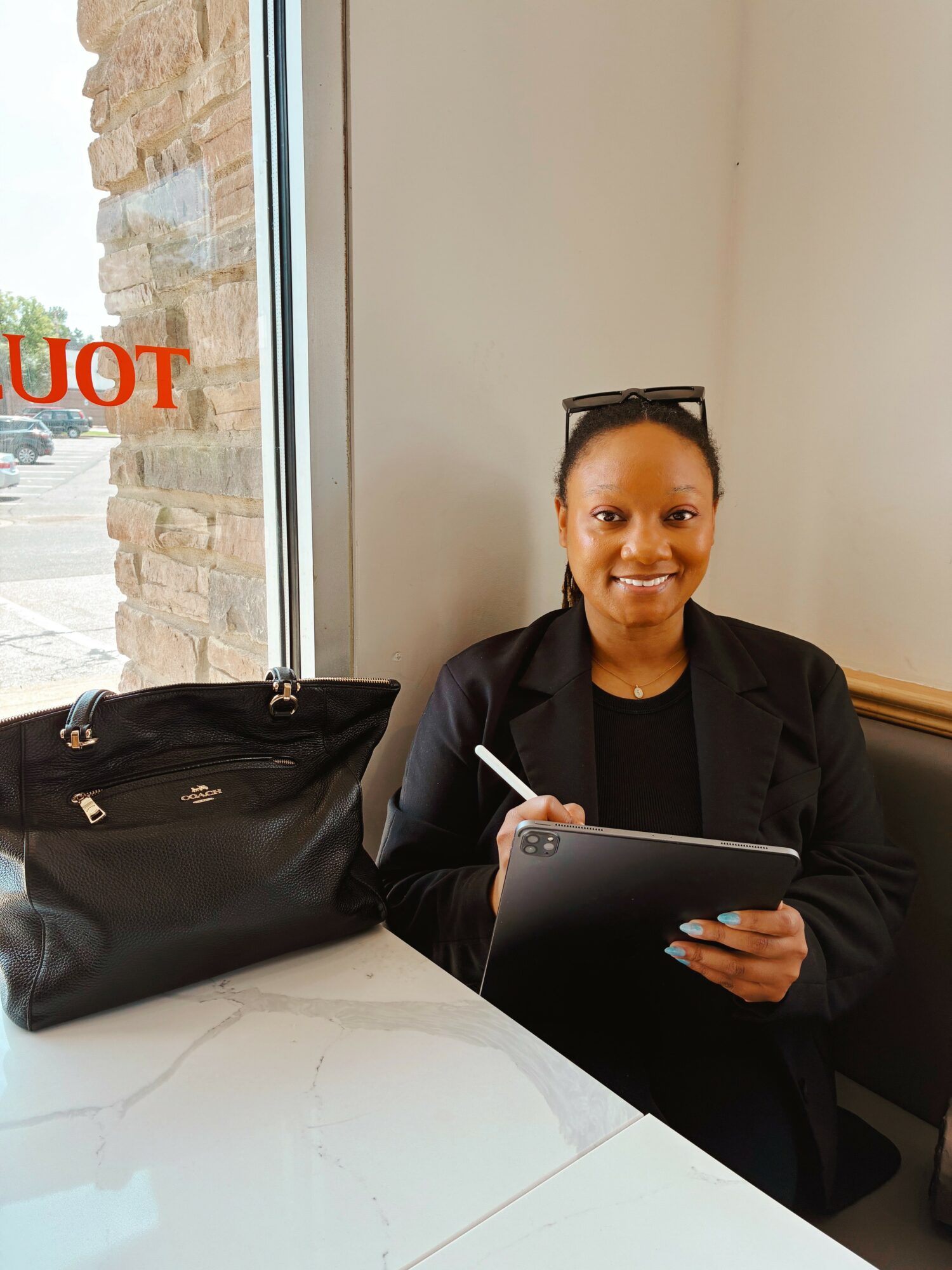 Woman sitting at a table with a tablet, smiling, with a black bag beside her, near a window.