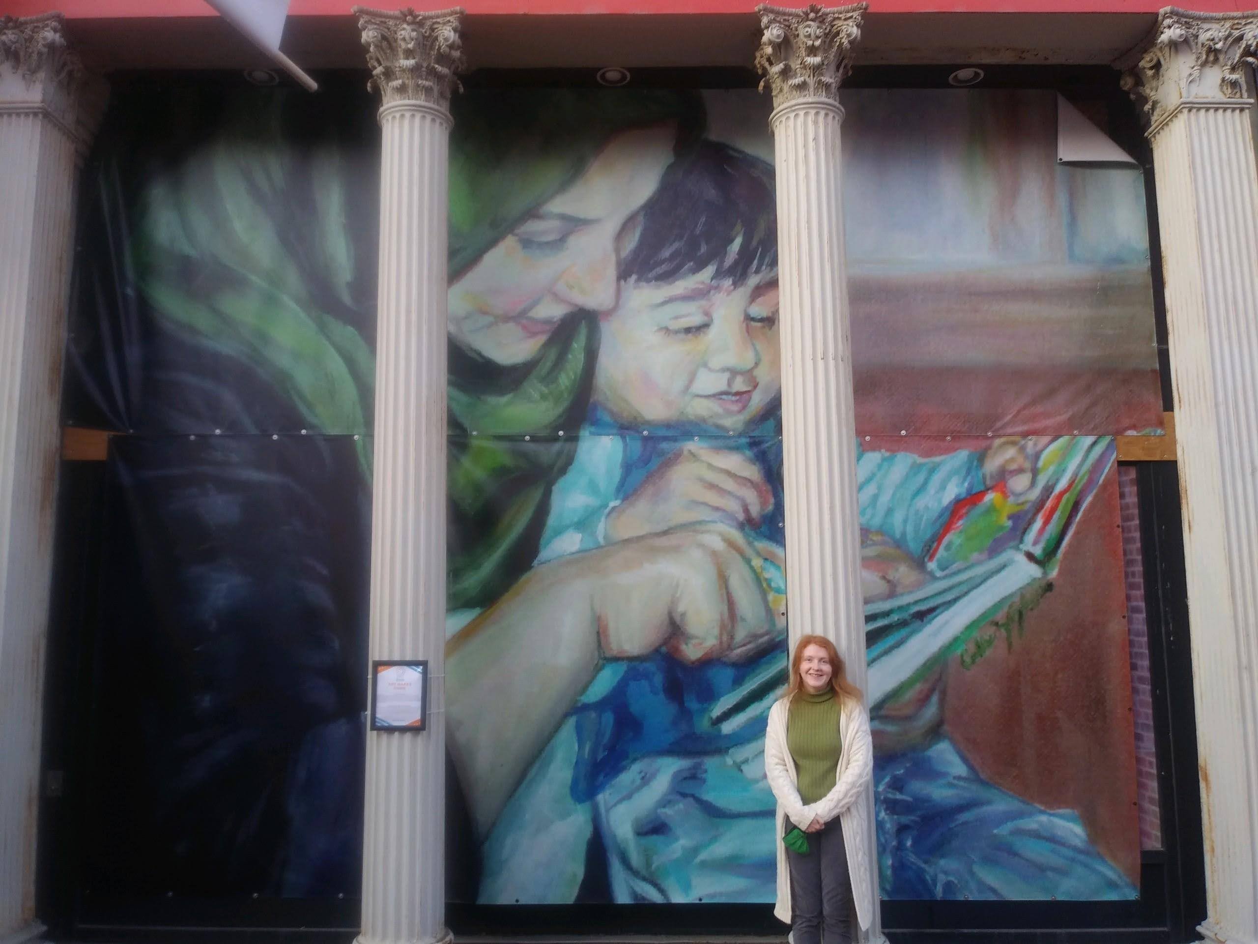 A woman stands in front of a colorful mural of a woman and child reading a book, framed by four columns.