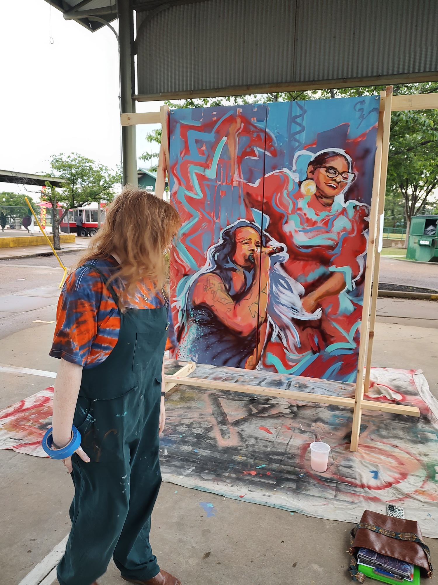 Person with curly hair wearing overalls and a colorful shirt looks at a mural of two women singing, with a dragon in the background.