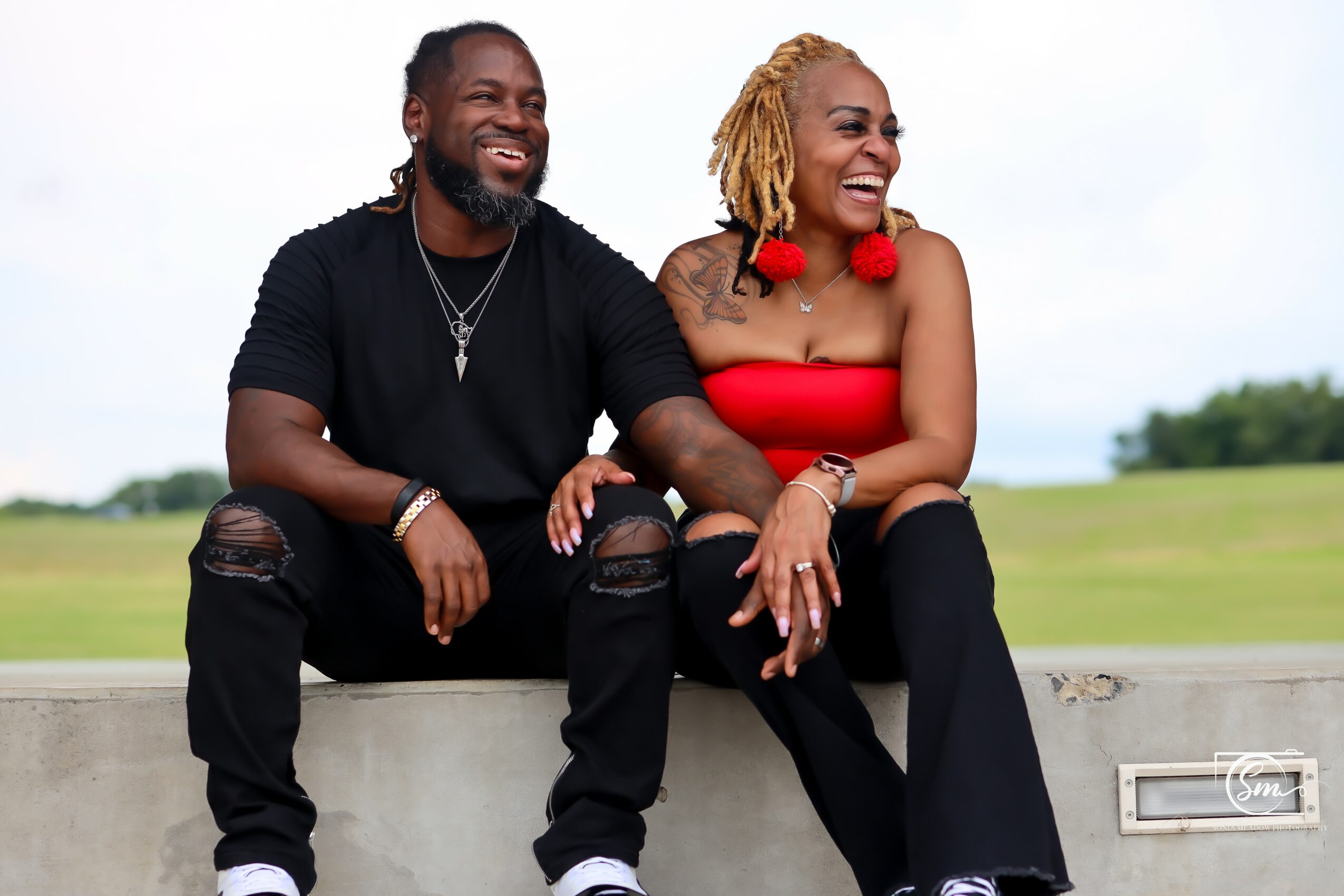 A smiling man and woman sit together outdoors on a concrete ledge, with a grassy field and sky in the background.