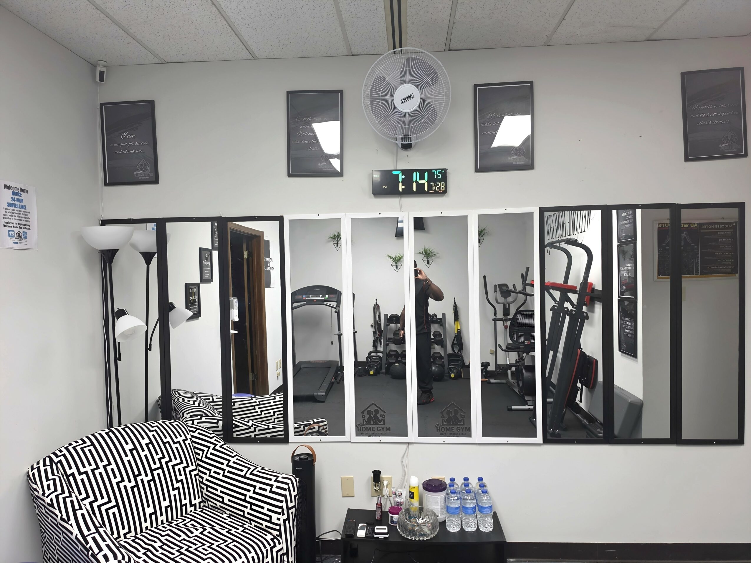 Gym room with mirrors, exercise equipment, and a person working out, viewed from a couch with patterned fabric.