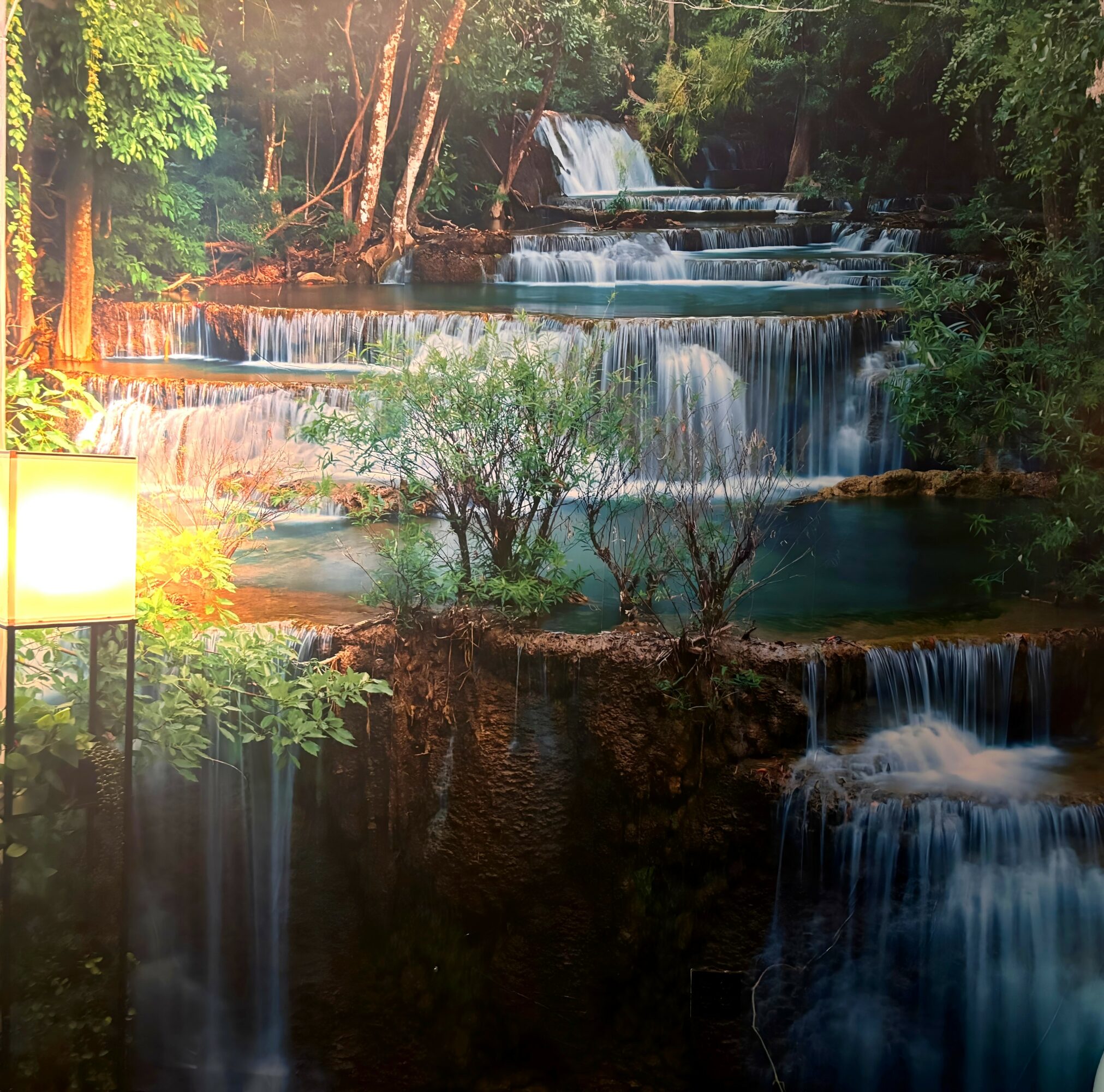 Indoor waterfall scene with multiple tiers of water flowing over rocks, surrounded by trees and a lamp on the left.