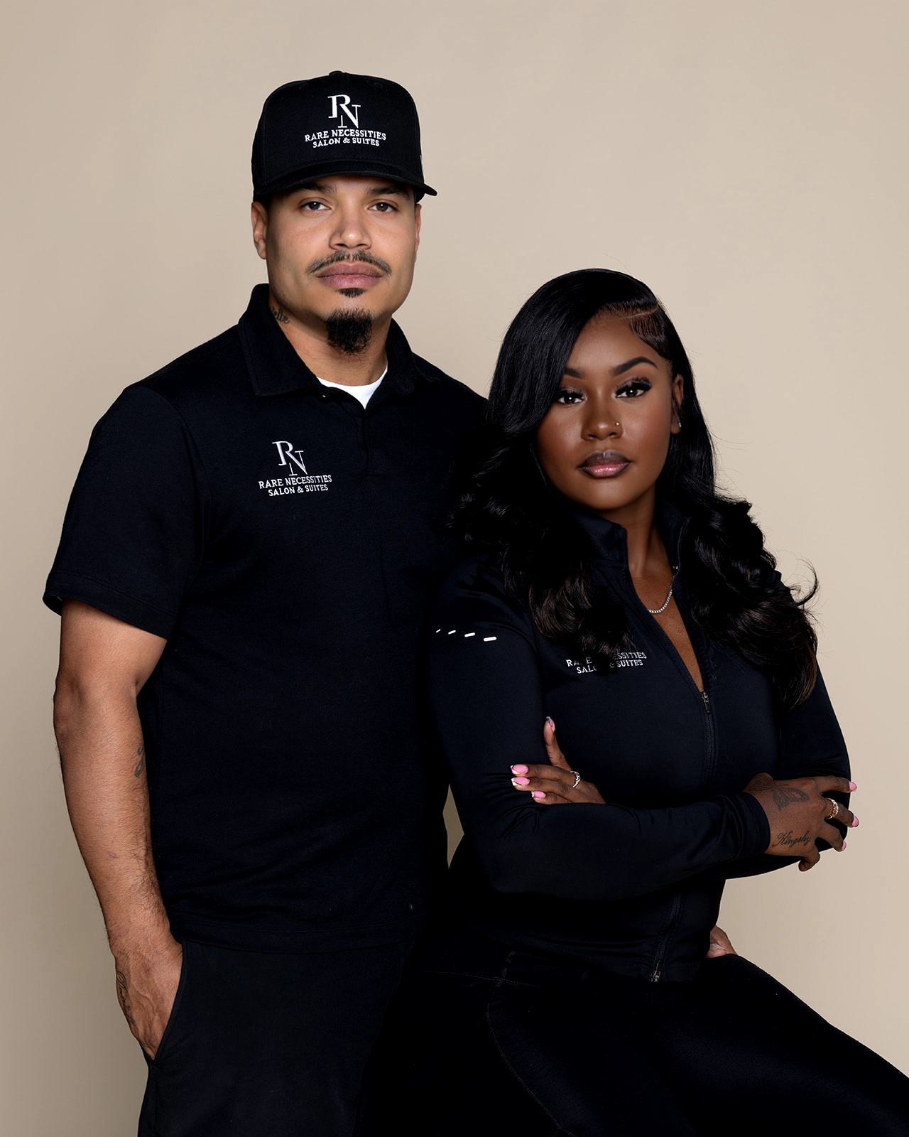 Two people, a man and a woman, stand side by side against a beige background, both wearing black clothing and caps.