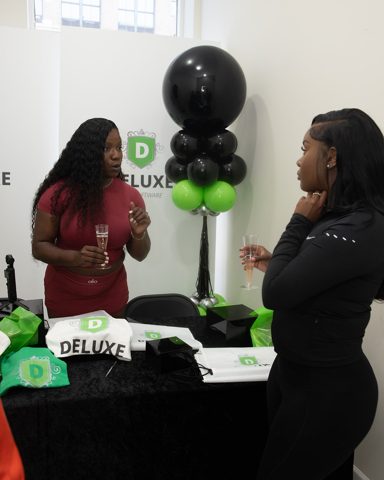 Two women talking at a table with a black and green balloon arrangement behind them, holding glasses, in a white room.