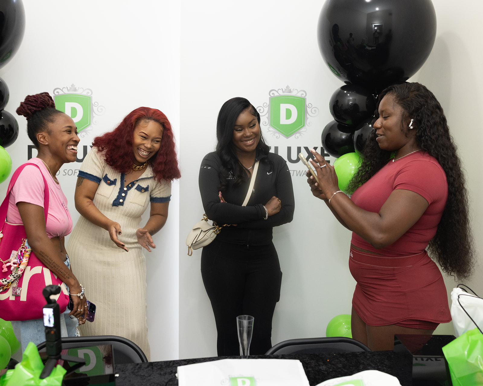 Four women standing and smiling indoors, with balloons and a table in front, one woman taking a photo.