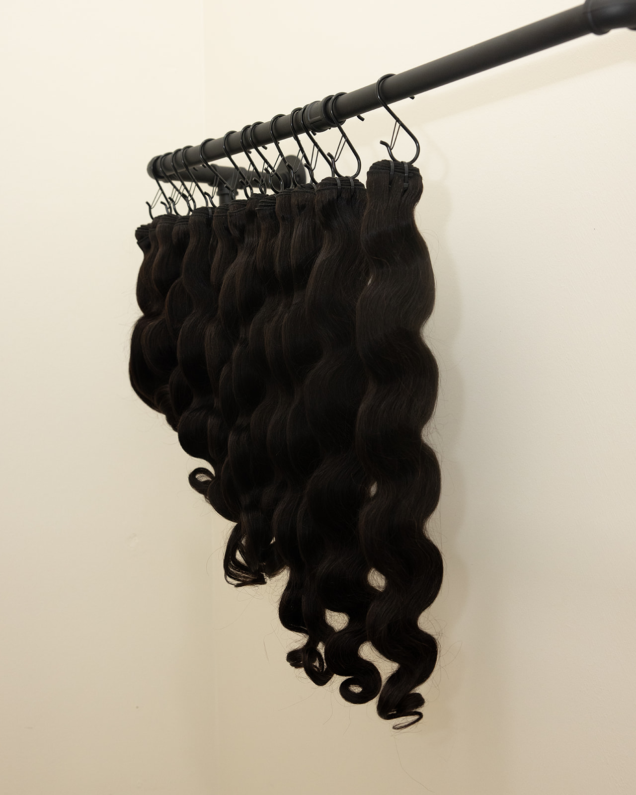 Multiple black curly hair extensions hanging from a black rod against a white wall.