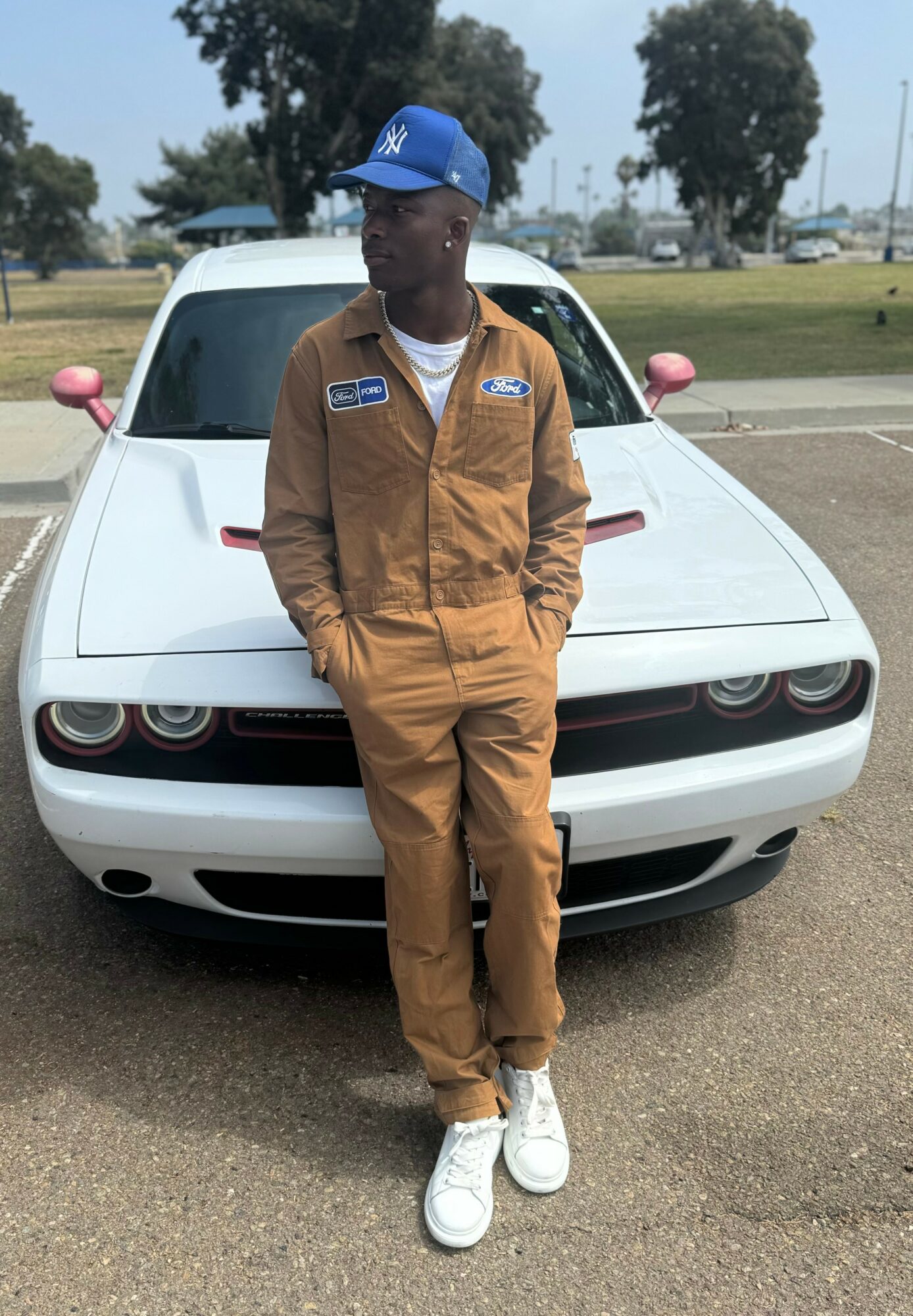 Person in brown jumpsuit and blue cap standing in front of a white sports car outdoors, with trees and sky in background.