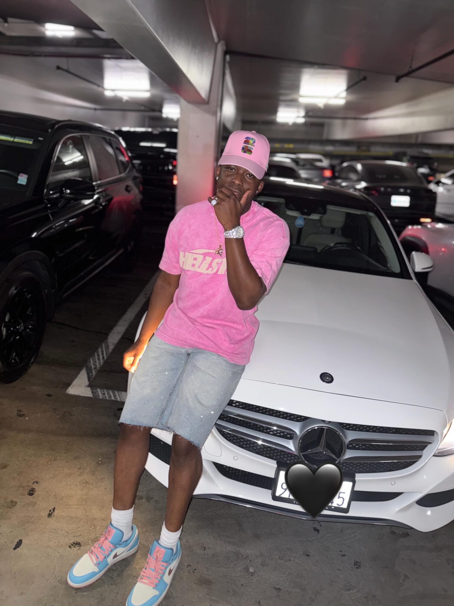 Person wearing pink cap and shirt, gray shorts, standing in parking garage next to white car, covering face with hand.