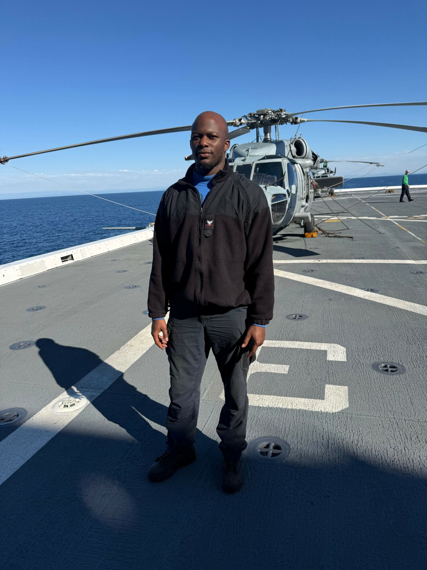 Man standing on a boat deck with boats and water in the background, wearing a black jacket and pants, facing left.