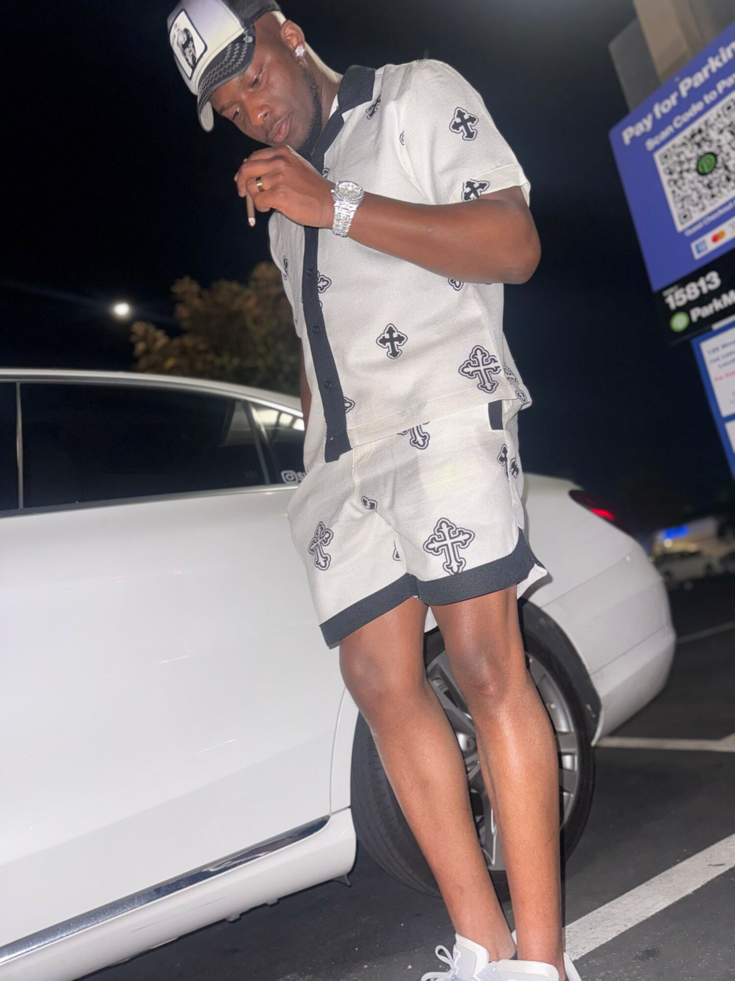 Young man in white patterned outfit standing next to a white car at night, looking at his phone.