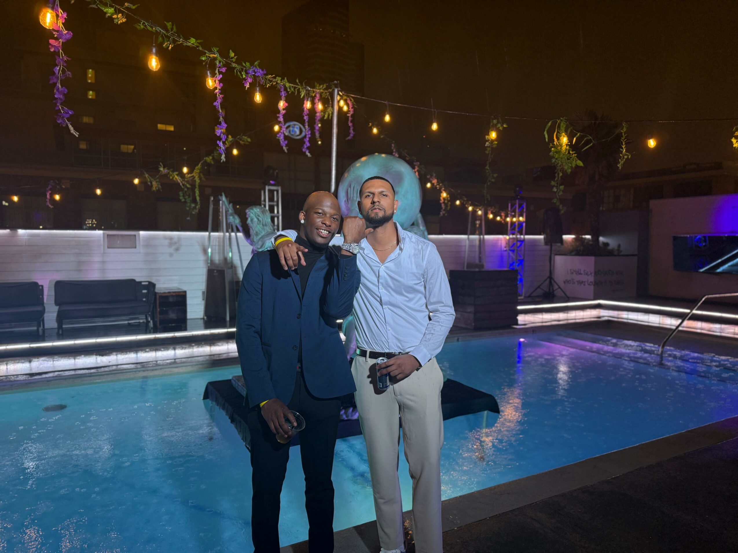 Two men stand by a pool at night, one with an arm around the other, decorated with string lights and hanging flowers.