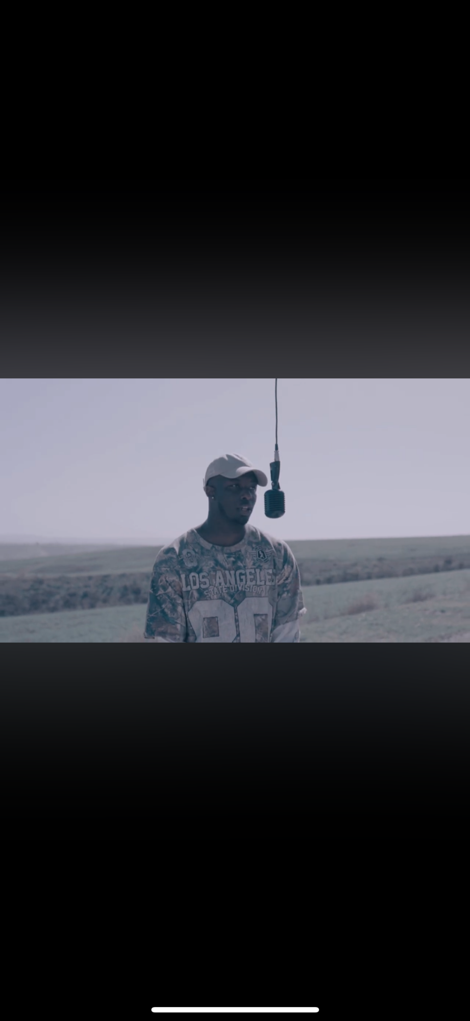 Person standing outdoors with a microphone hanging from above, landscape in background, wearing a patterned shirt and cap.