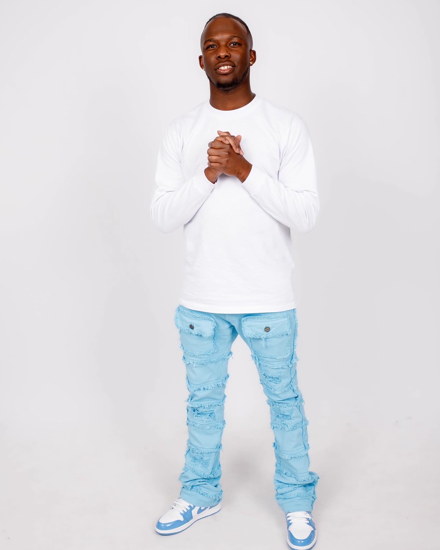 Man standing with hands clasped, wearing a white long-sleeve shirt and light blue distressed jeans, smiling, plain white background.