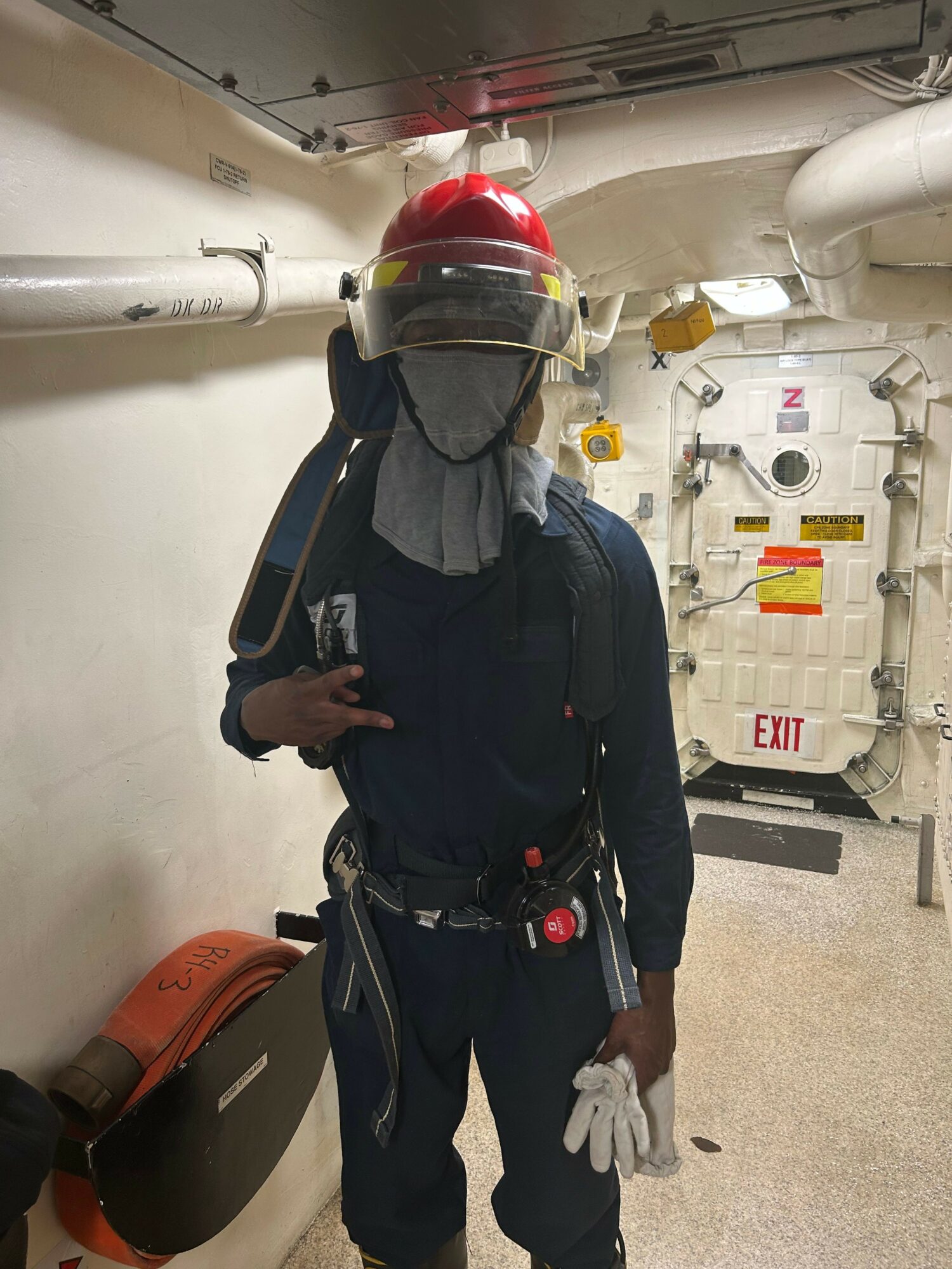 Person in uniform with helmet and gloves standing in a corridor, holding a phone, with equipment and signs in the background.