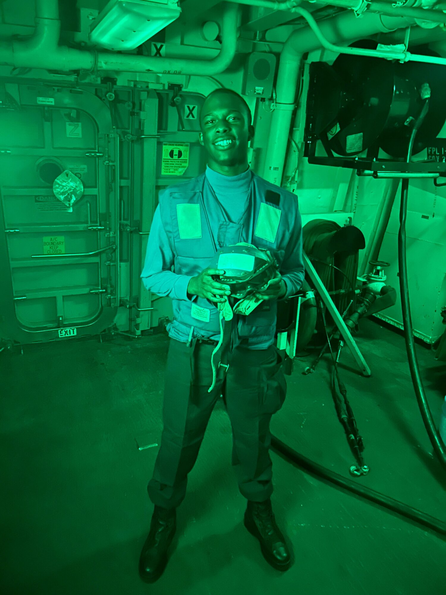 Person in a blue uniform with equipment, standing in a room with pipes and machinery, holding a phone, with green lighting.