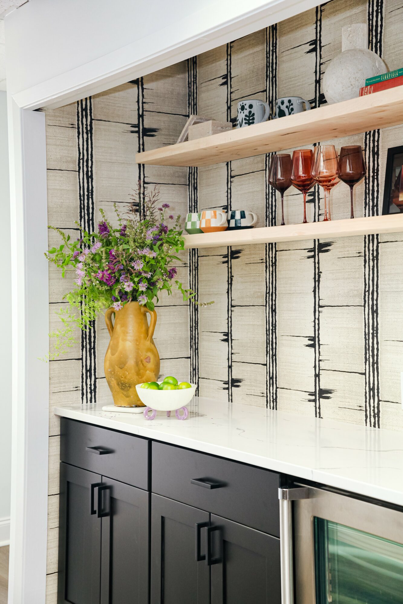 Shelf with glasses and books above a countertop, decorated with a vase of flowers and a bowl, against a patterned wall.
